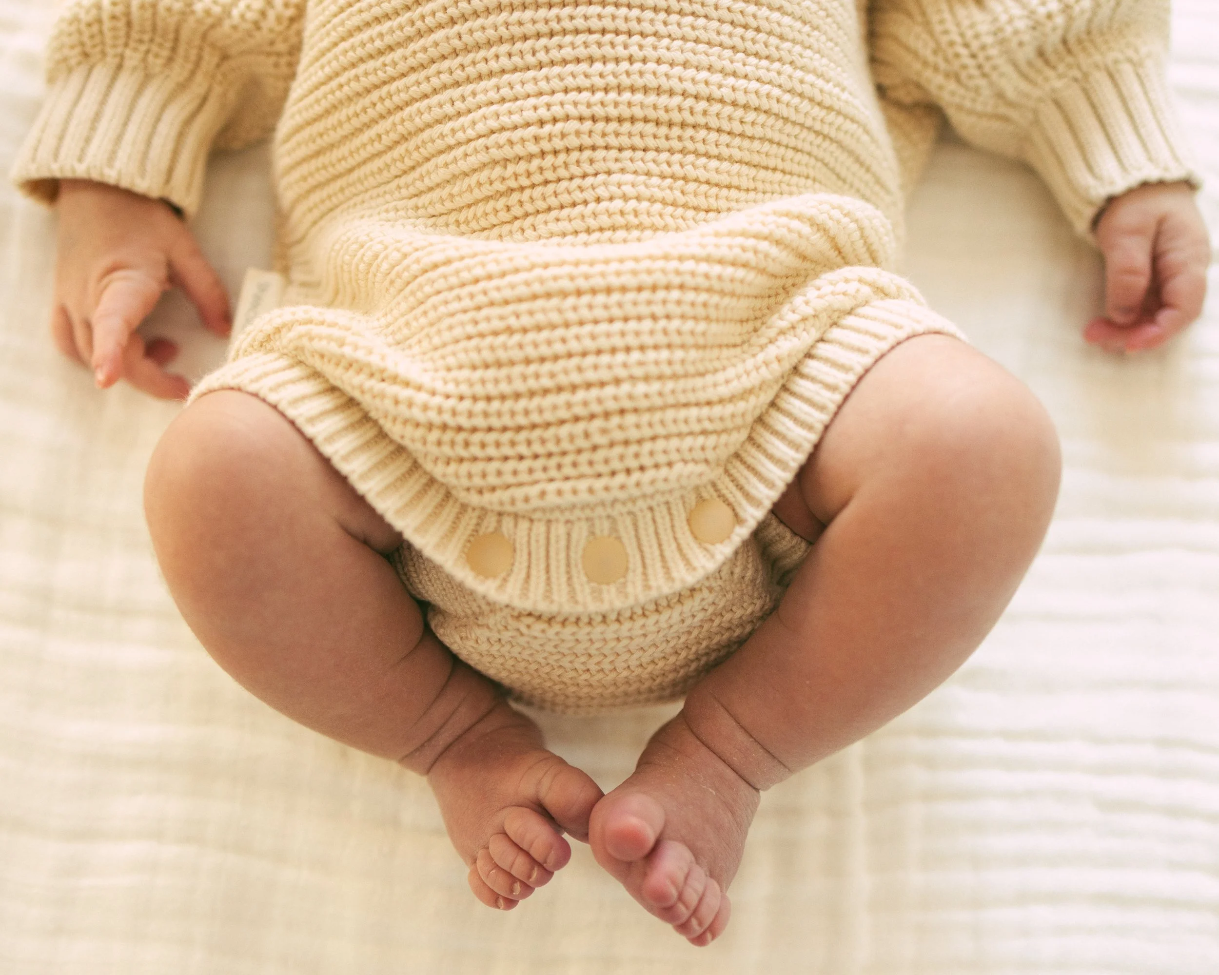 Close-up of a baby's hand gripping an adult's finger while sitting on a beige blanket, wearing a beige knit sweater.