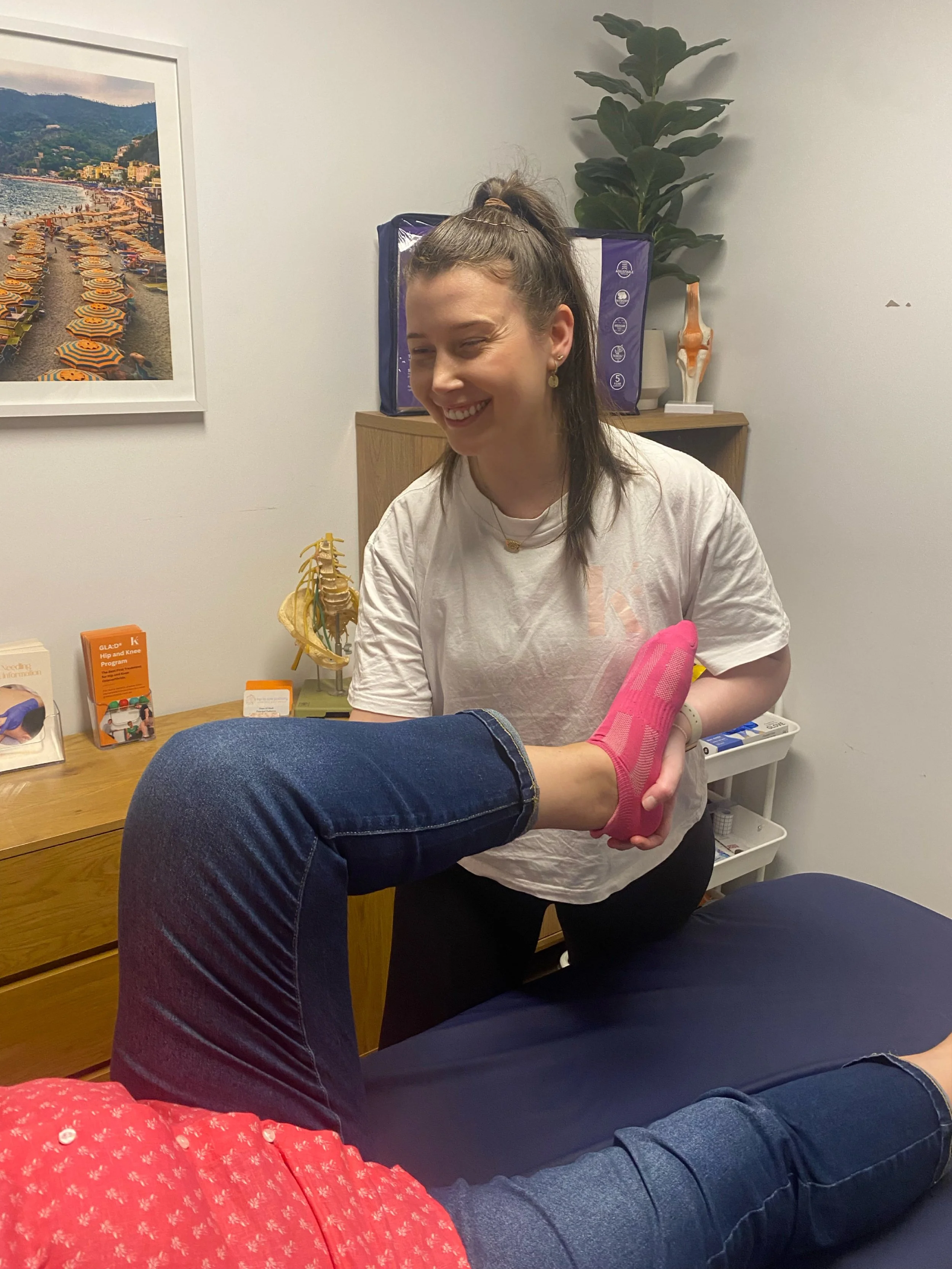 A woman in a white t-shirt is smiling and holding a person's foot with a pink sock, during a physical therapy session in an office with medical models and a plant in the background.