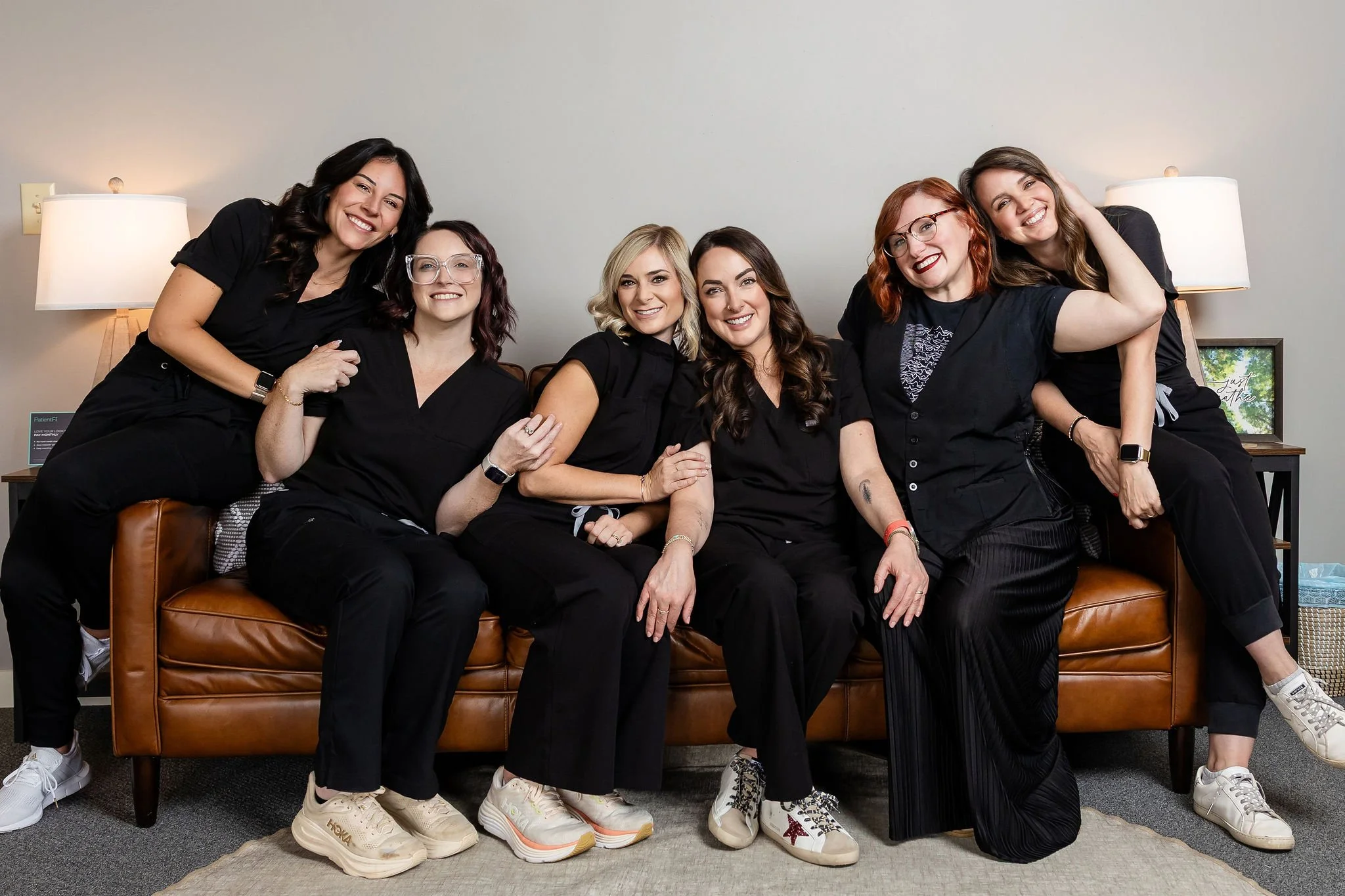 Group of six women wearing black clothing, sitting and standing on a brown leather couch in a cozy room, smiling and posing for a photo.