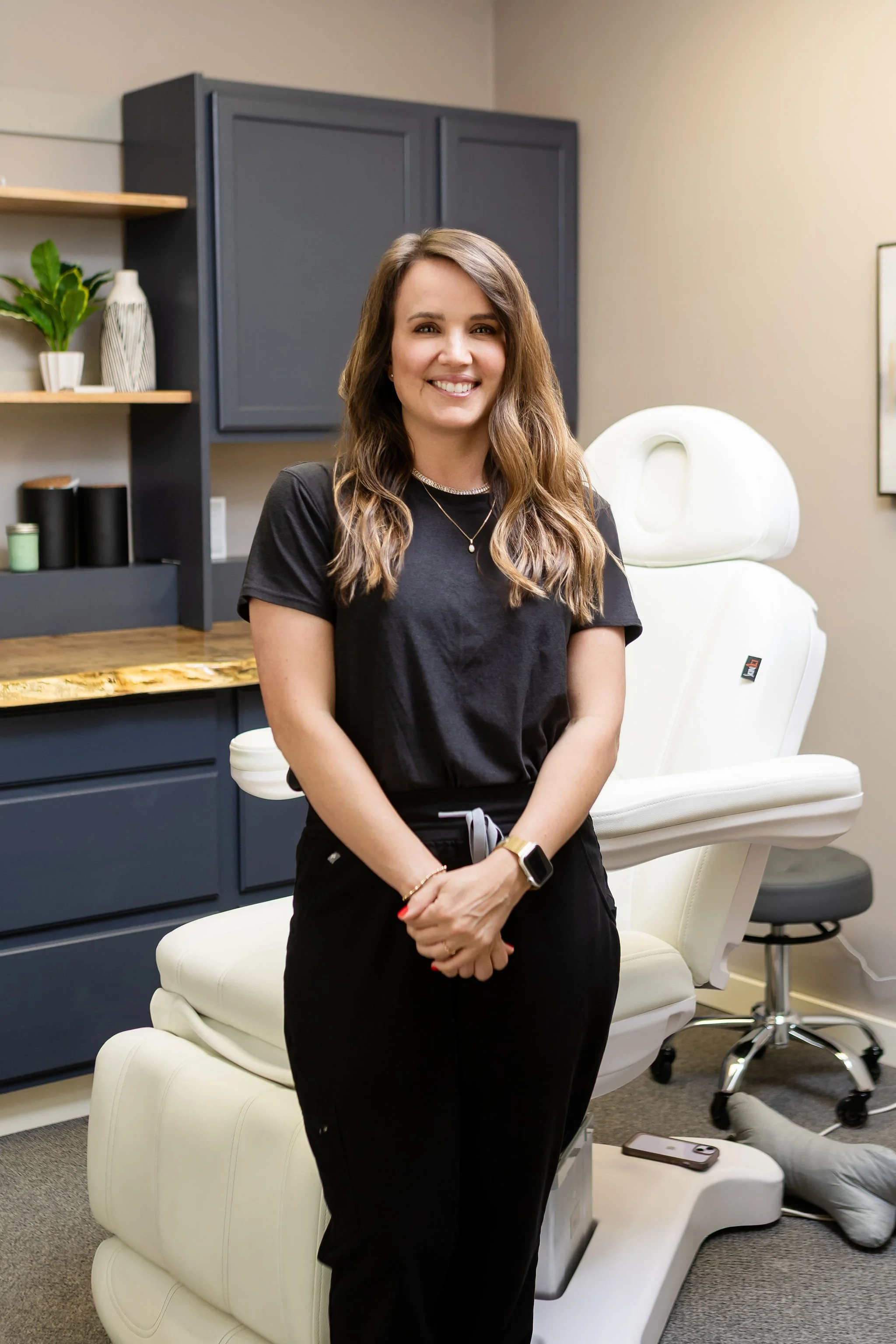 A woman standing in a medical office with a massage or examination chair behind her, smiling at the camera.