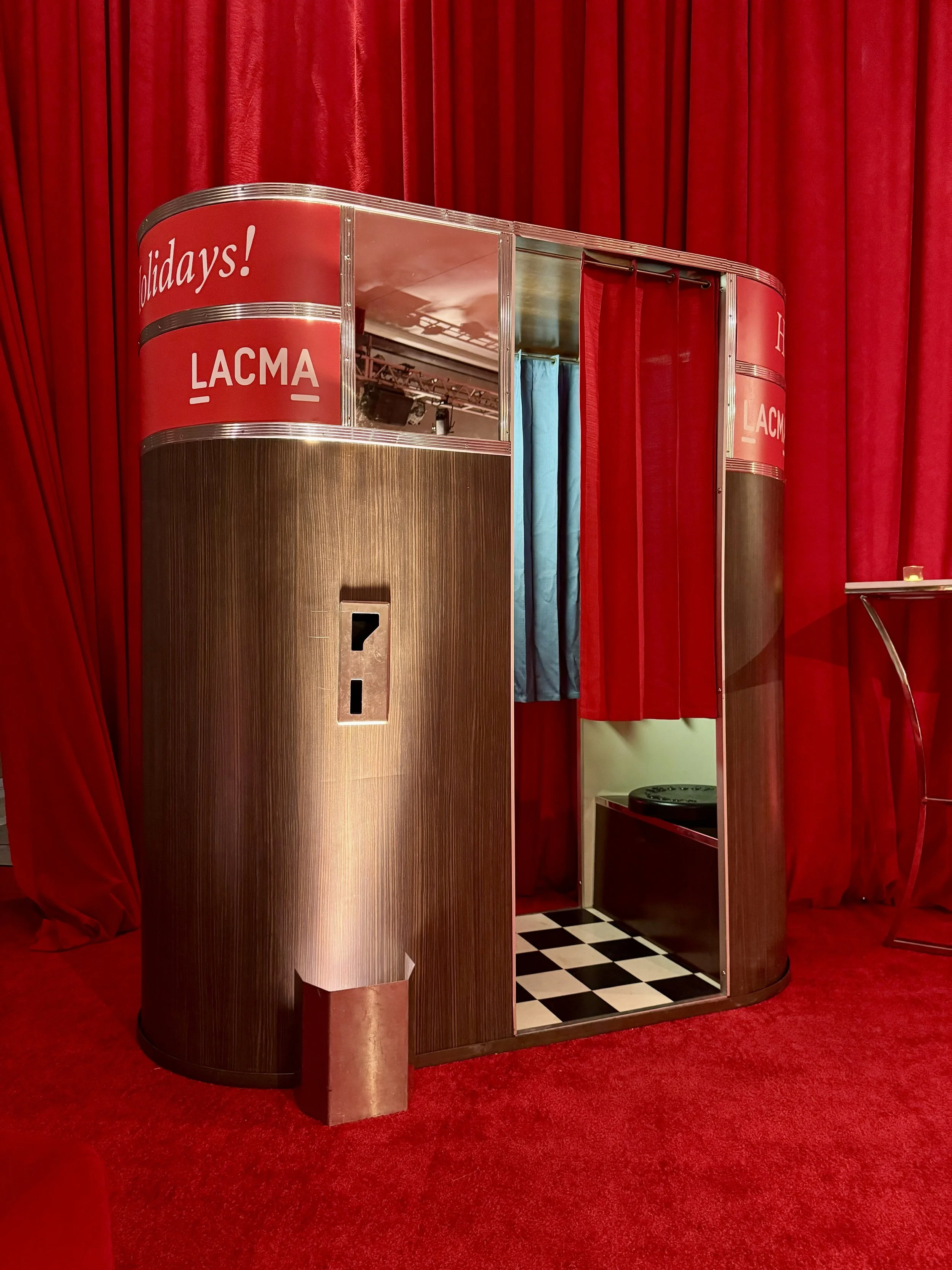 A vintage analog photo booth with a red front curtain at the LACMA with a LACMA decal on it and a red curtain background and red floor. Model 11