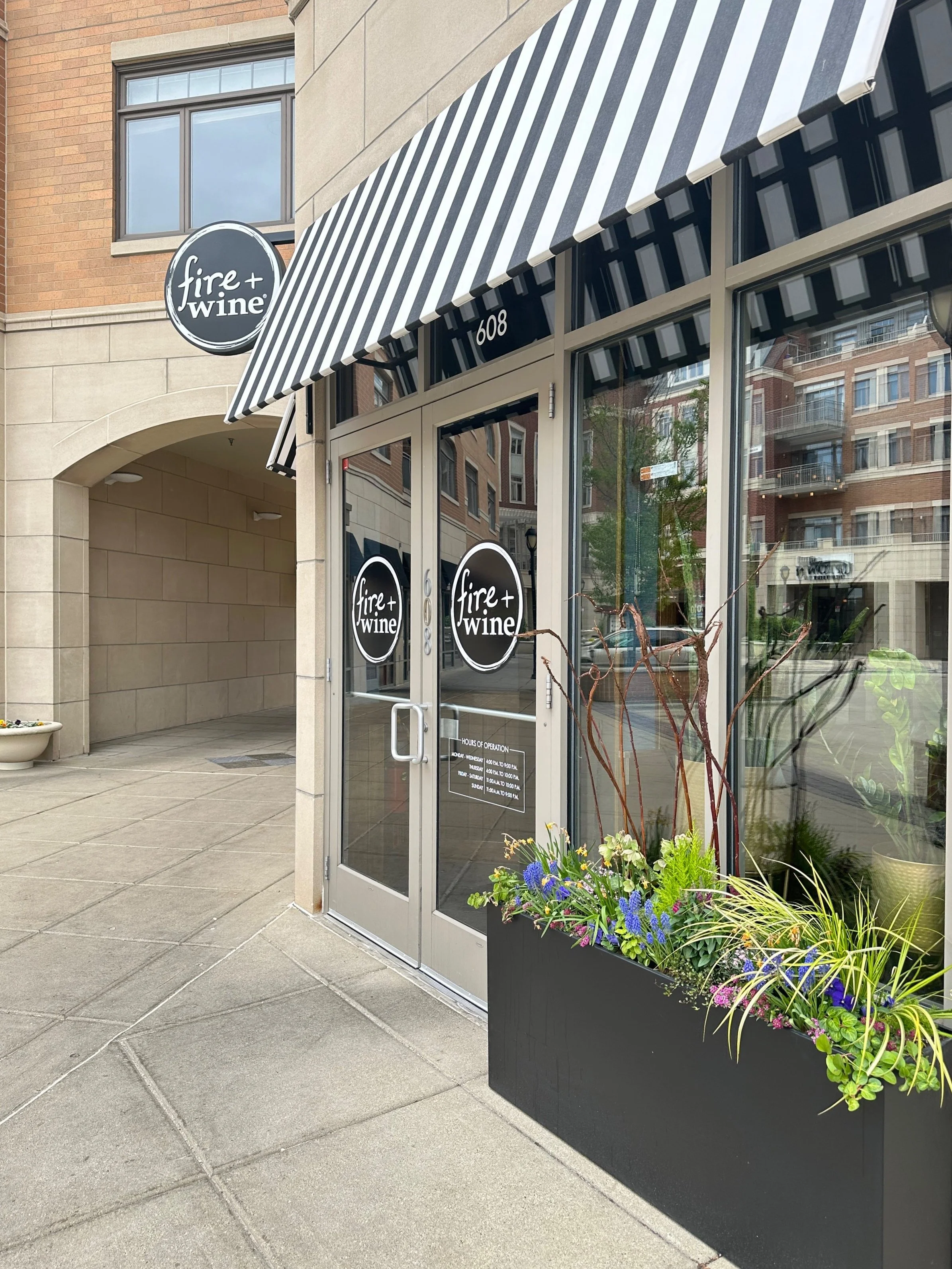 Storefront with striped awning labeled 'fire + wine', with glass doors and a black planter box with purple and green plants outside.