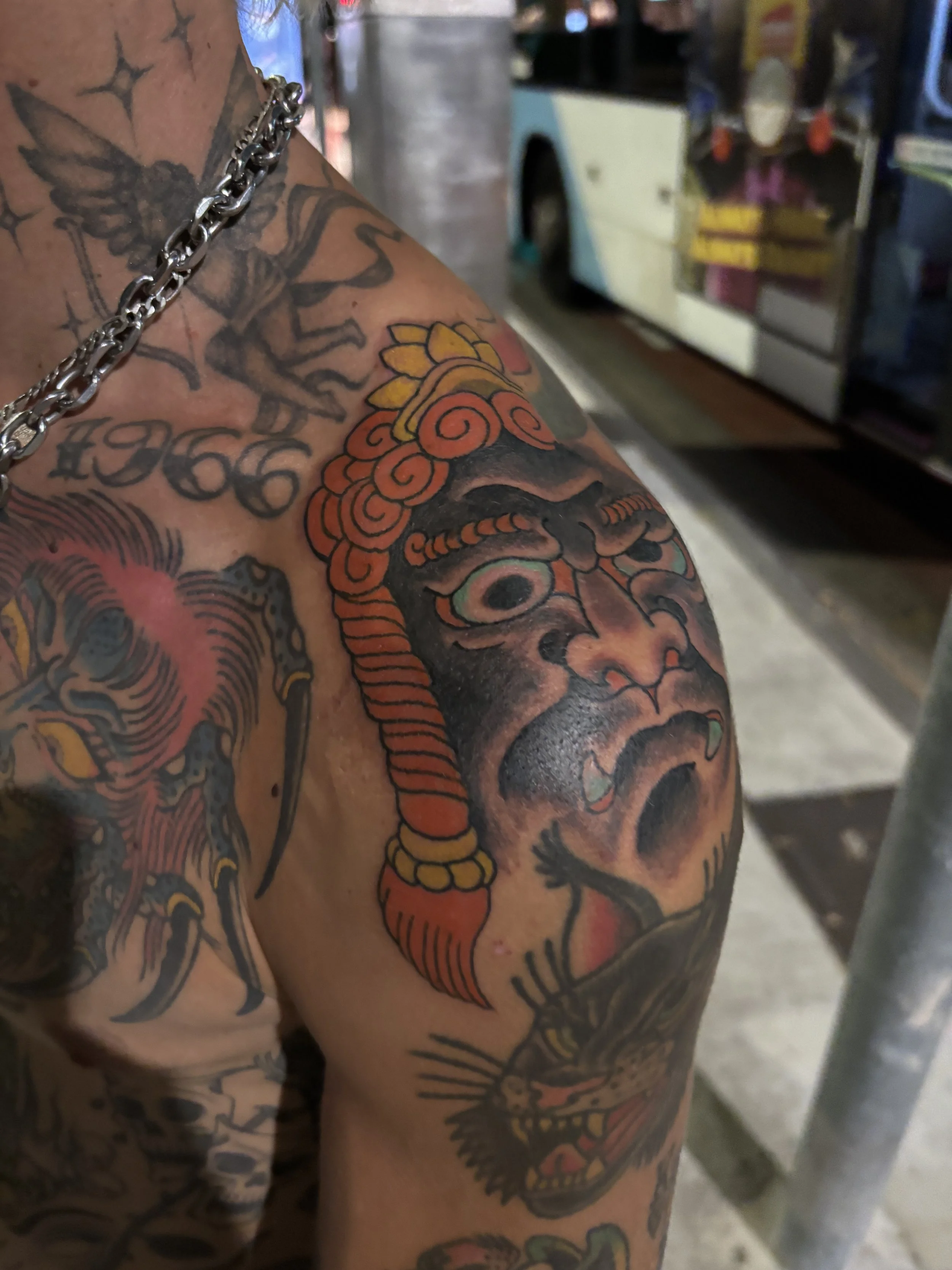 Close-up of a person's arm and shoulder covered in colorful tattoos, including a prominent face of a woman with framed hair, with a dragon and other designs visible in the background. The person is wearing a silver chain necklace.