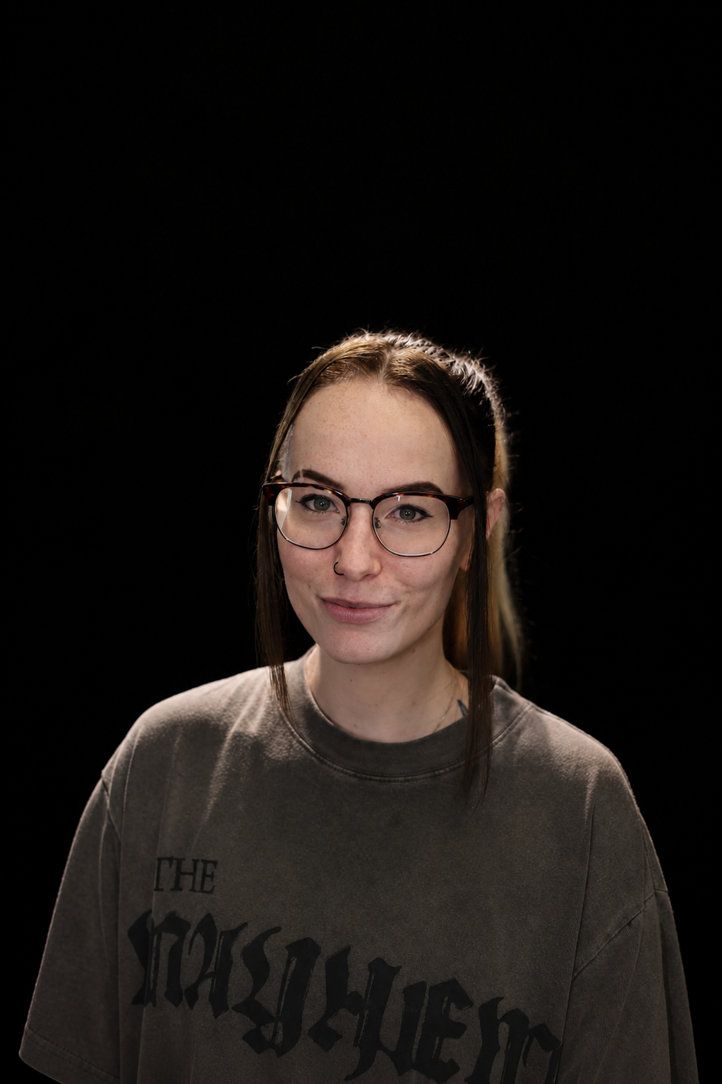 Portrait of a young woman with glasses and a nose ring, wearing a gray T-shirt, standing against a black background.