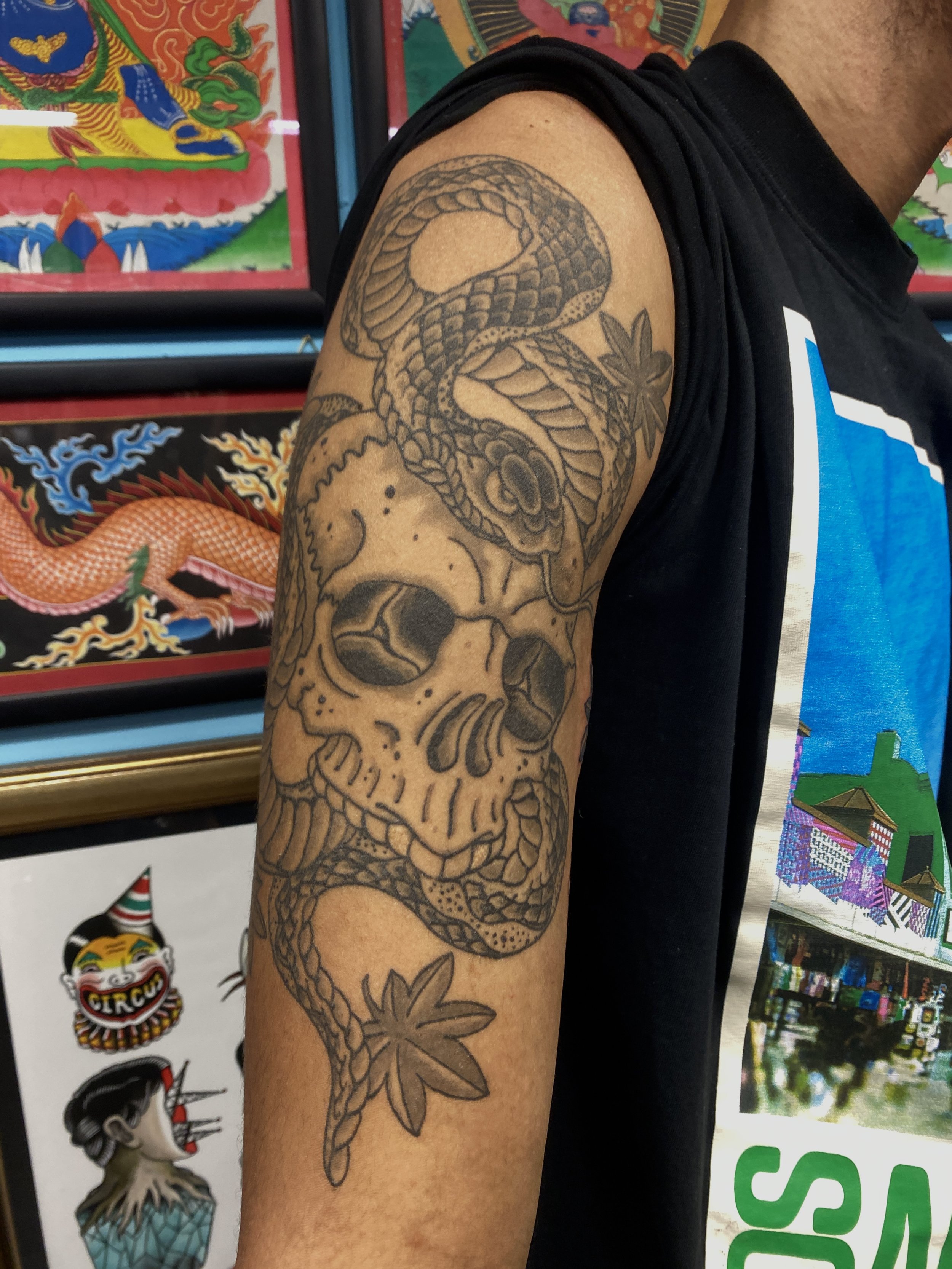 A person's arm with a detailed black and gray tattoo of a snake entwined around a skull and a flower. The background includes colorful artwork and stickers.