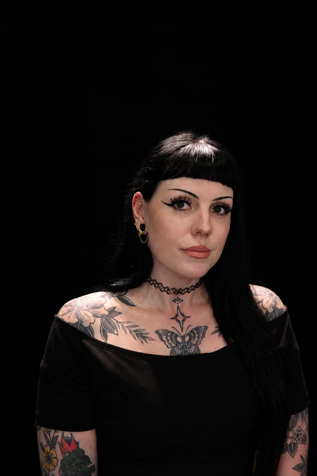 Portrait of a woman with black hair and tattoos on her shoulders and arms, wearing a black top and black choker, against a black background.