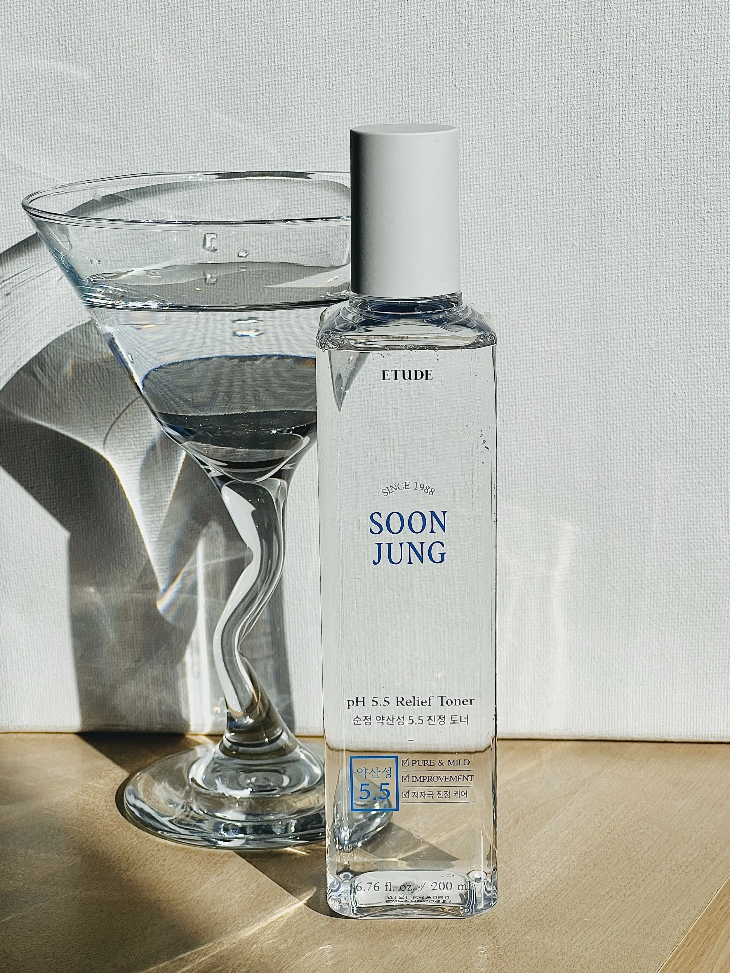 A clear glass with a twisted stem and a bottle of SOON JUNG pH 5.5 Relief Toner on a wooden surface, with sunlight and shadows creating reflections on the glass and bottle.