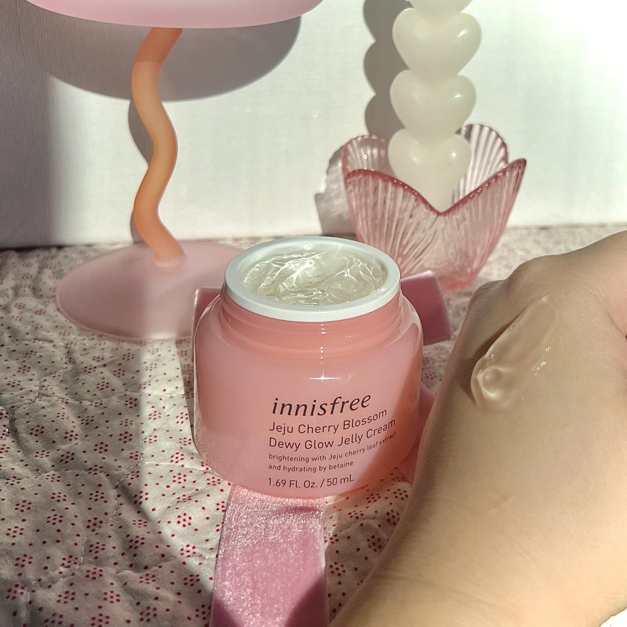 Open pink jar of Innisfree Jeju Cherry Blossom Dewy Glow Jelly Cream on a pink surface with decorative pink and white objects in the background.