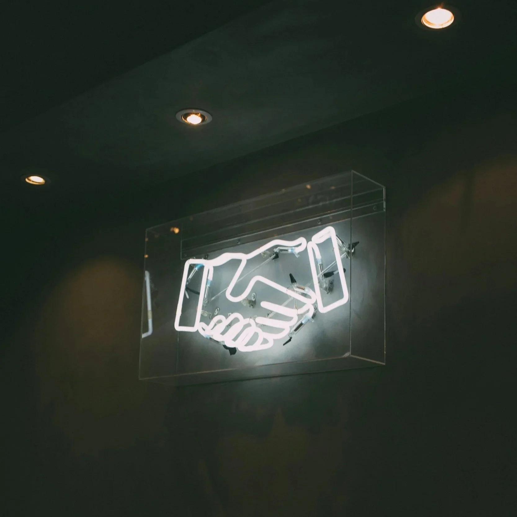 Neon sign depicting a handshake, mounted on a dark wall in a glass case, with overhead spotlights.