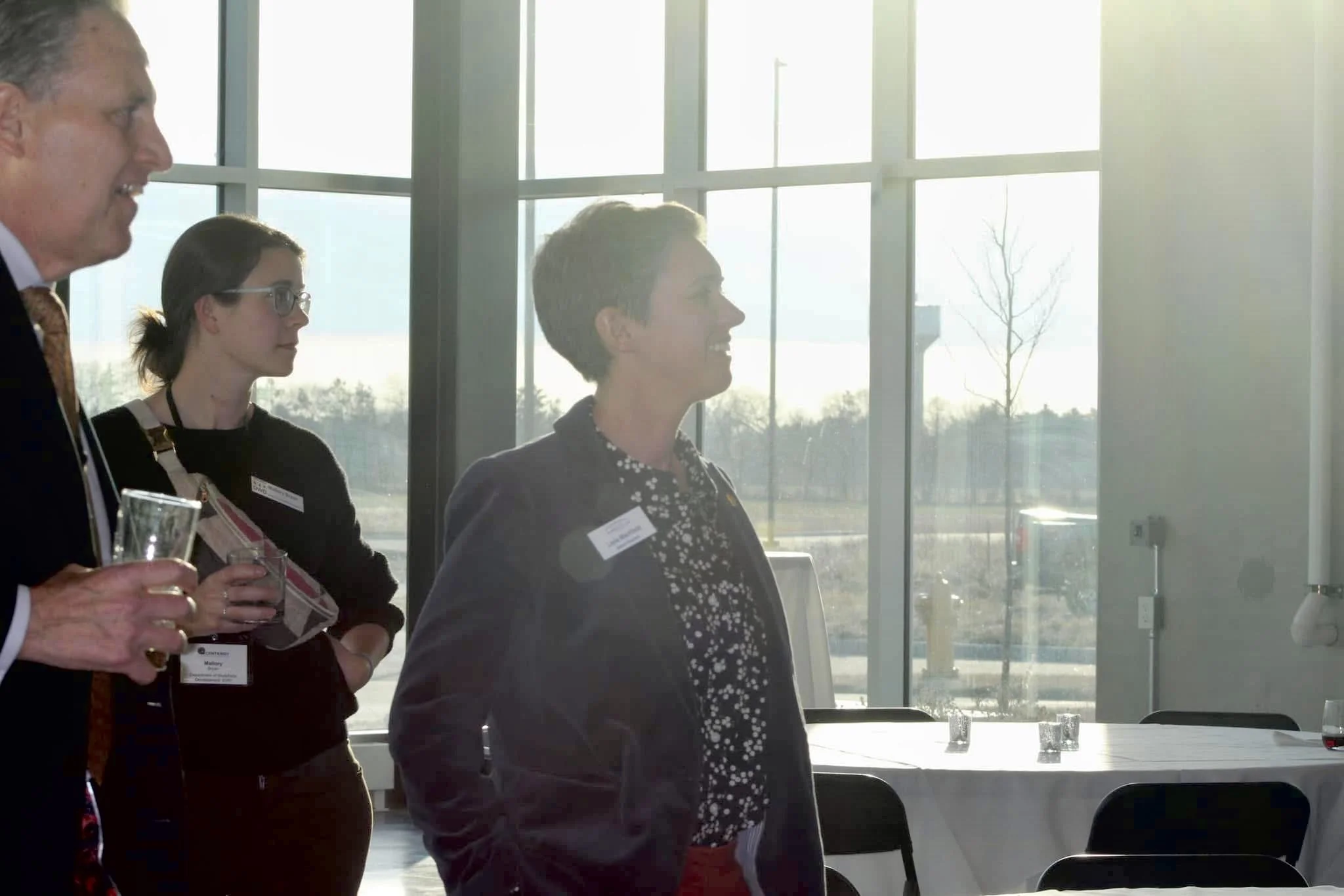 Three people standing in a room with large windows, having a conversation.