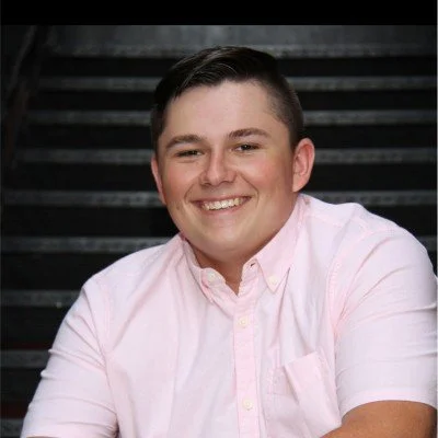 A young man with short dark hair smiling, wearing a light pink button-up shirt, posed against a dark background.