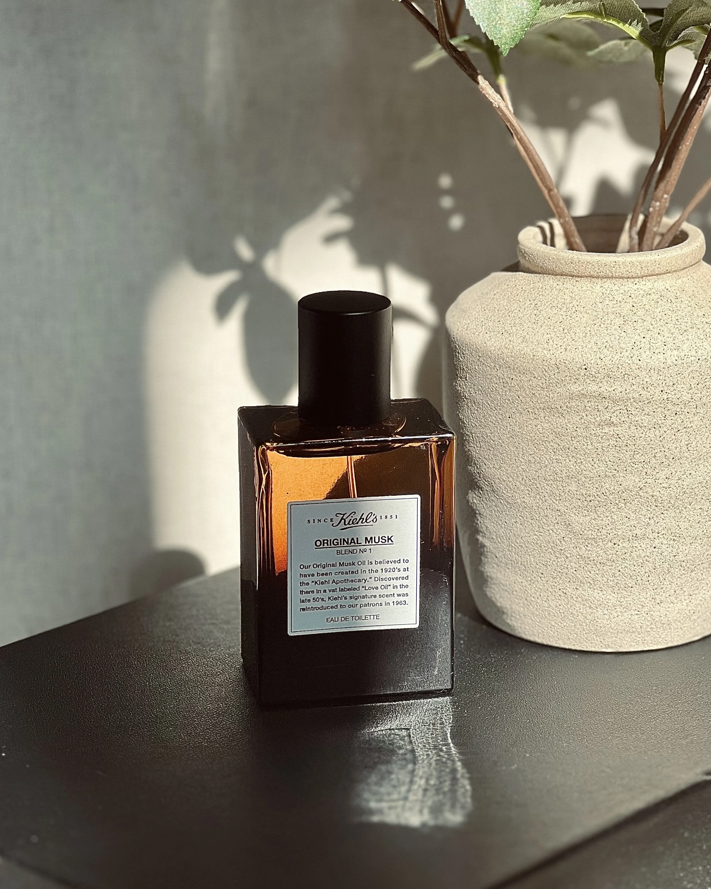 A bottle of Kiehl's Original Musk perfume next to a beige ceramic vase with green leaves and branches, with sunlight casting shadows on the wall behind.