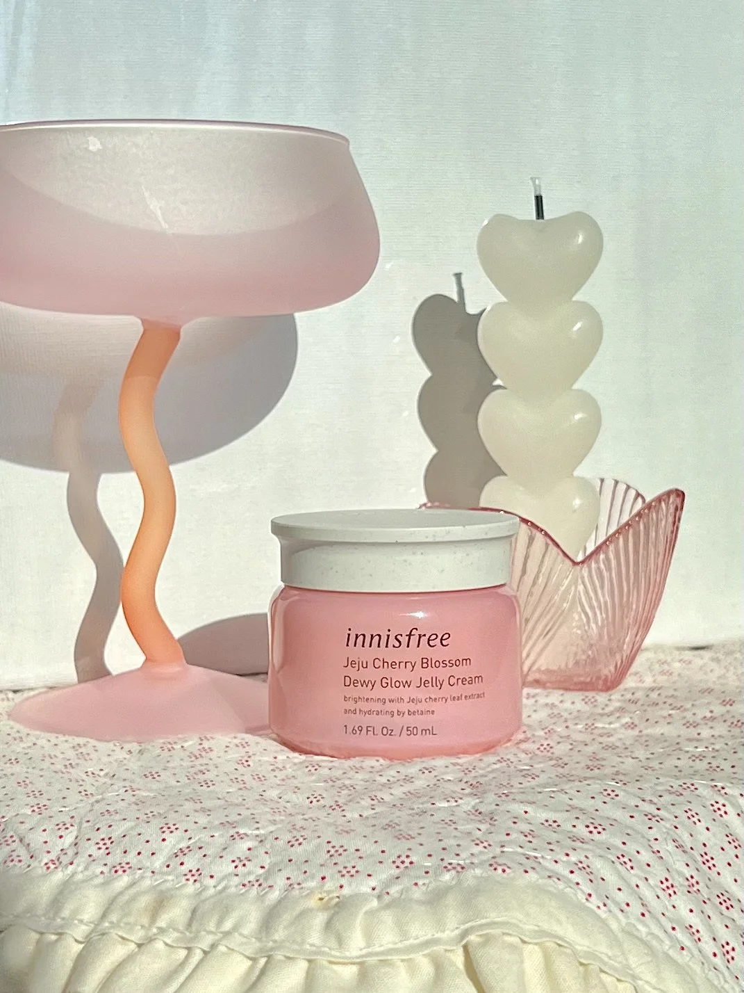Pink jar of Innisfree Jeju Cherry Blossom Dewy Glow Jelly Cream placed on a white lace tablecloth with pink floral pattern, surrounded by pink and white decorative objects including a large pink bowl with a wavy stand, a white bubble candle with a wick on top, and a pink glass bowl with a ruffled edge.