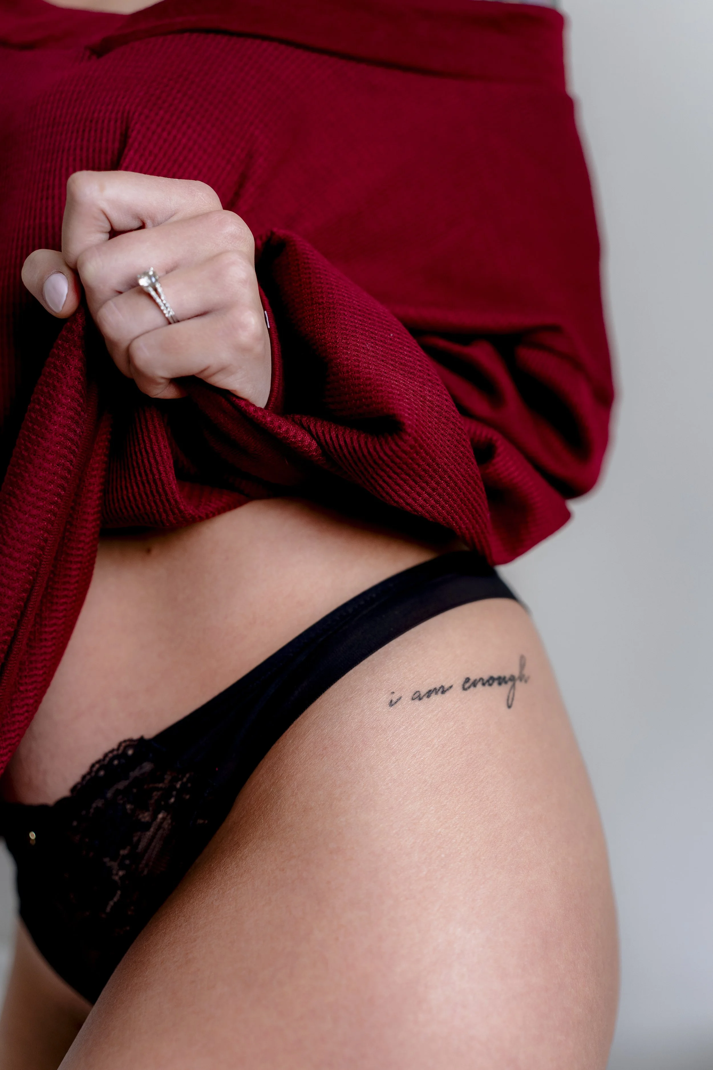A woman pulling up a red shirt to reveal a black lace underwear and a tattoo on her hip that says 'I am enough'.