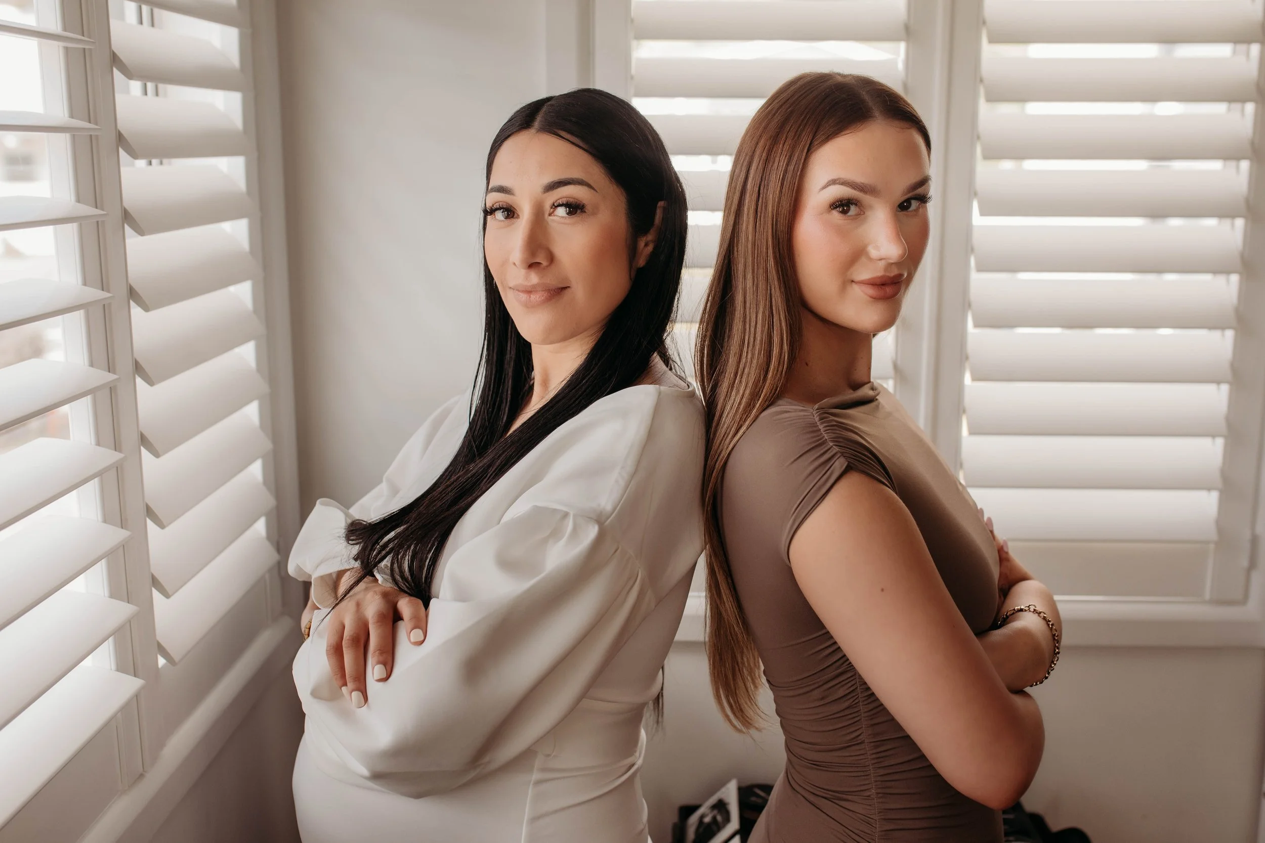Two women standing back-to-back with arms crossed, smiling, in front of white window shutters.
