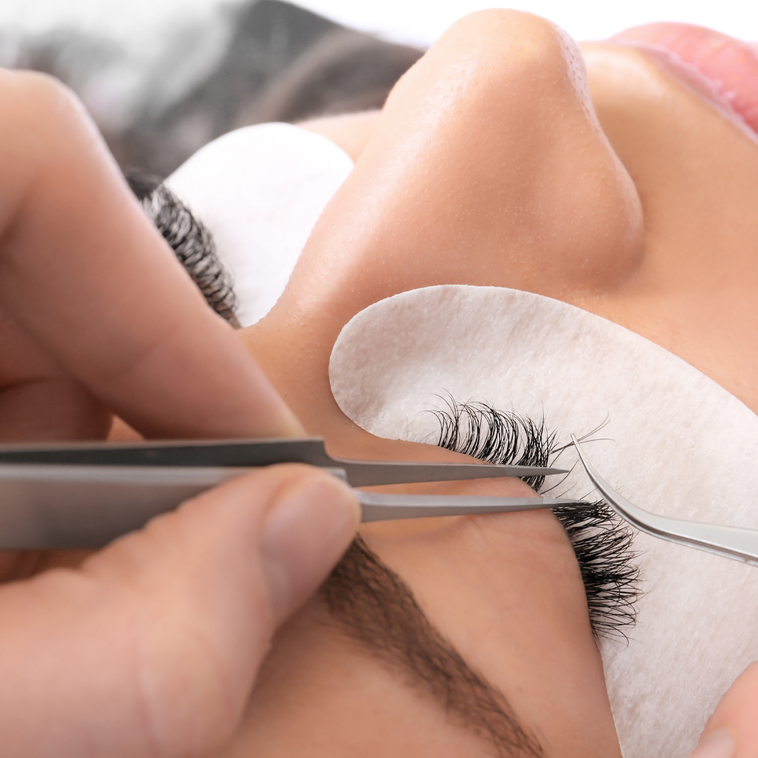 Close-up of a person receiving eyelash extension treatment, with an aesthetician using tweezers to apply individual false eyelashes while a cotton pad holds natural lashes down.