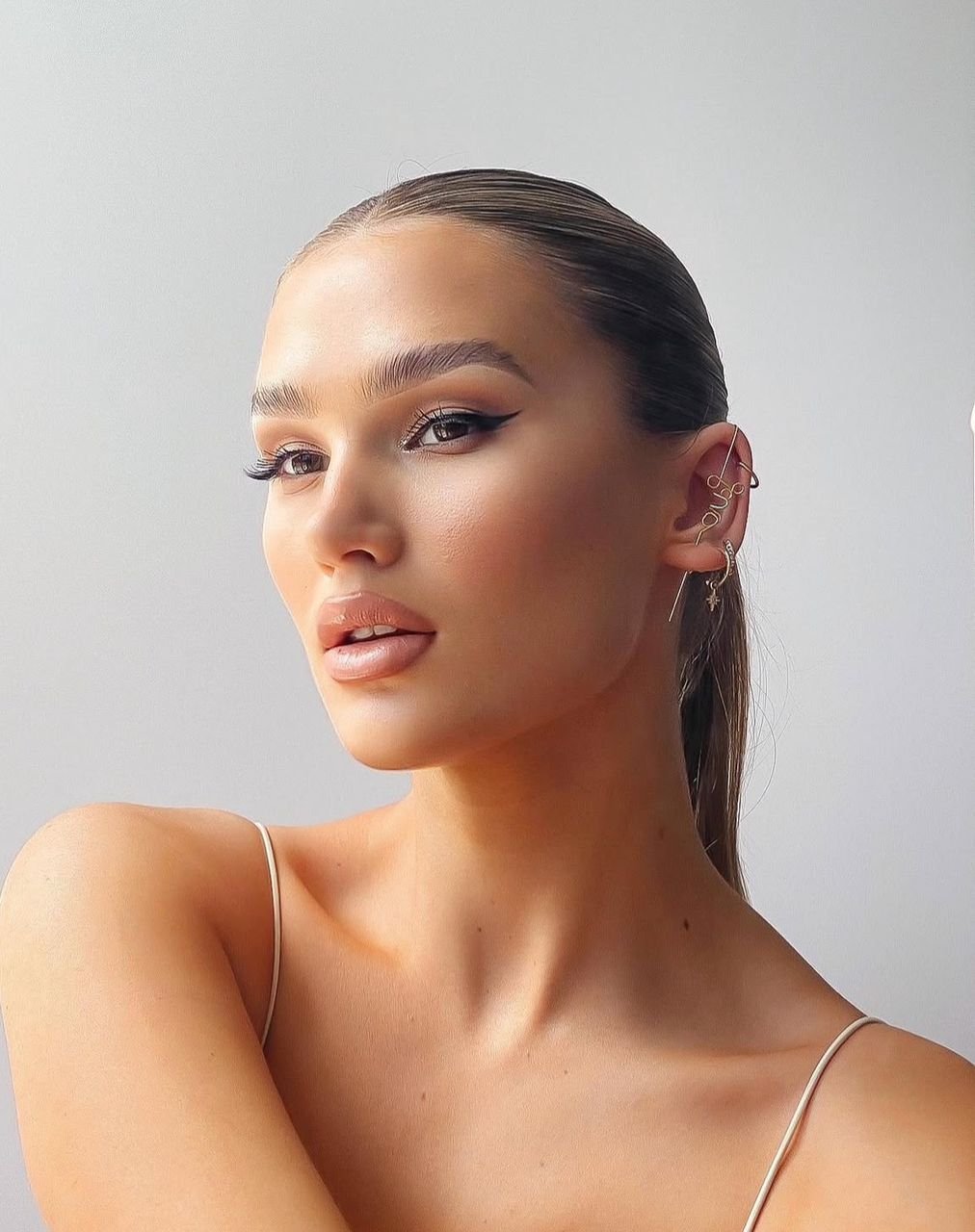 Close-up of a young woman with slicked-back hair, winged eyeliner, and hoop earrings, wearing a spaghetti strap top.