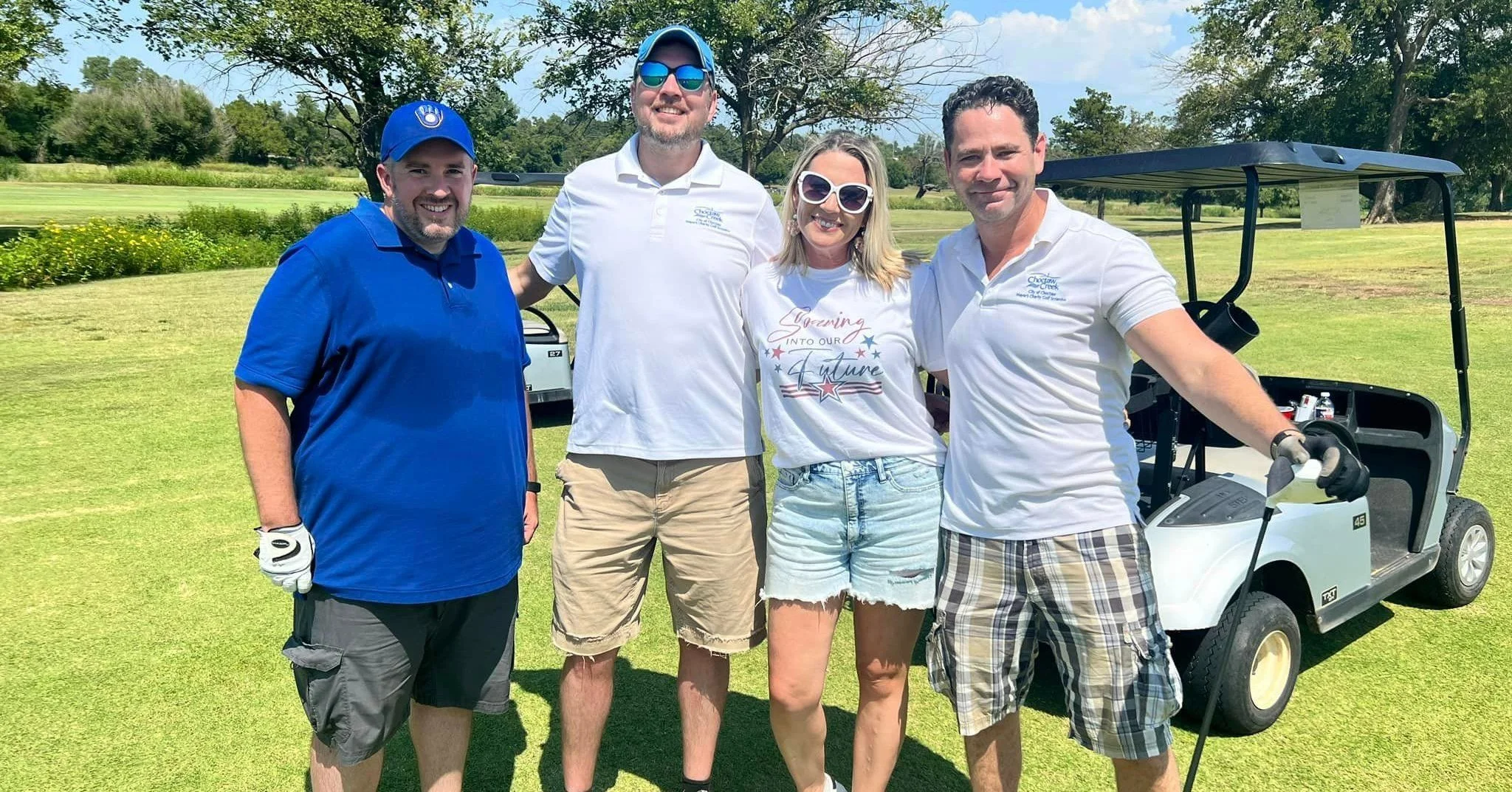 Four people standing on a golf course smiling for the camera, with a golf cart in the background.