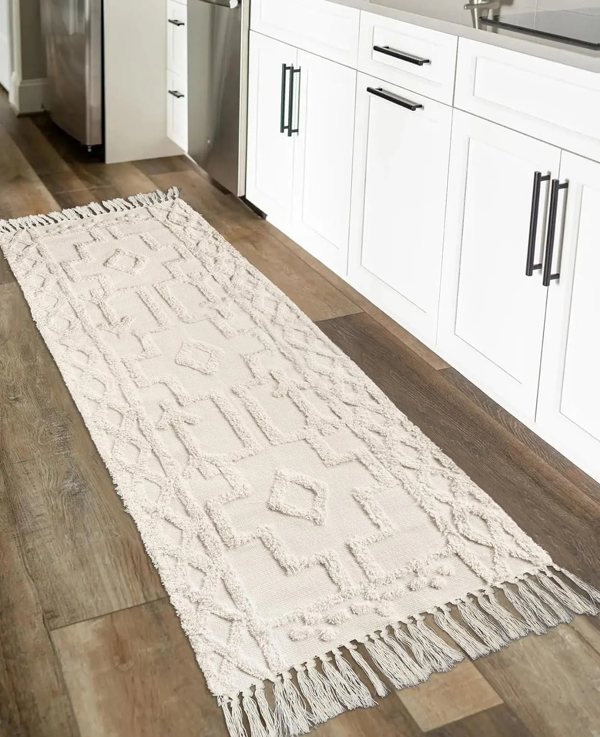 A white textured rug with fringe on a hardwood kitchen floor, next to white cabinets with black handles.