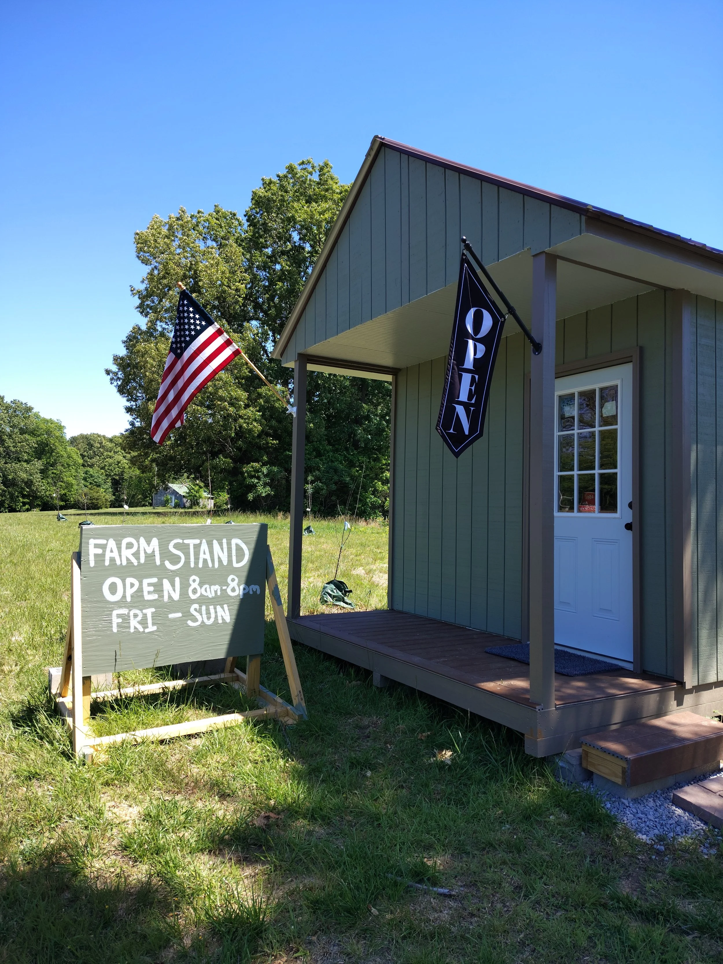 A small gray farm stand with a porch, a door, an American flag, a black "Open" flag, and a handwritten sign on grass indicating the farm stand is open from 8 am to 8 pm Friday to Sunday. Trees and a clear blue sky are in the background.