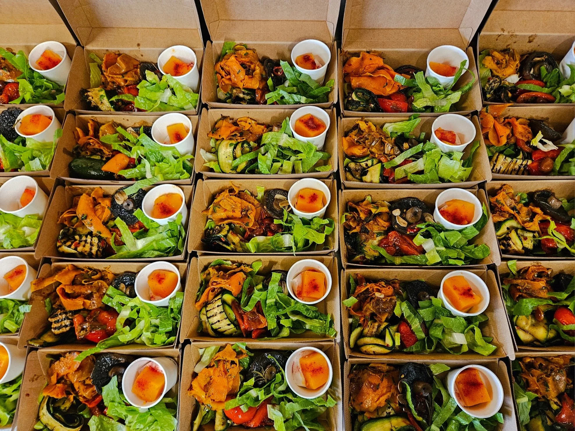 Multiple takeout bio food containers filled with various grilled and roasted vegetables, topped with fresh lettuce, and small sweet treat