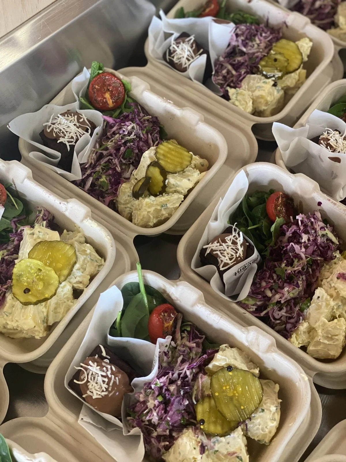 Multiple takeaway trays with salads, pickles, mashed potatoes, and chocolate desserts.
