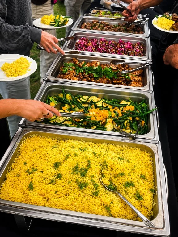 Buffet table with various dishes including saffron rice, grilled vegetables, different meat dishes, and colorful salads, with people serving themselves.