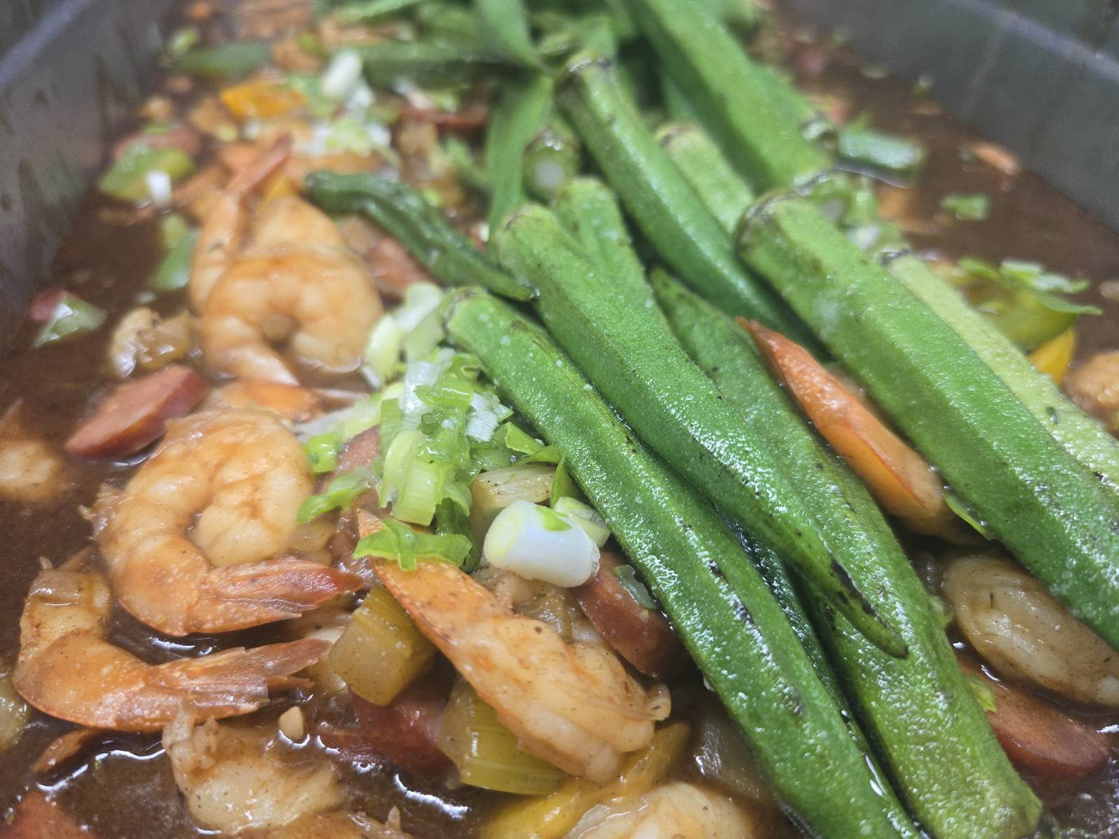 Close-up of a dish with cooked shrimp, chopped green onions, and okra in a brown sauce or broth.