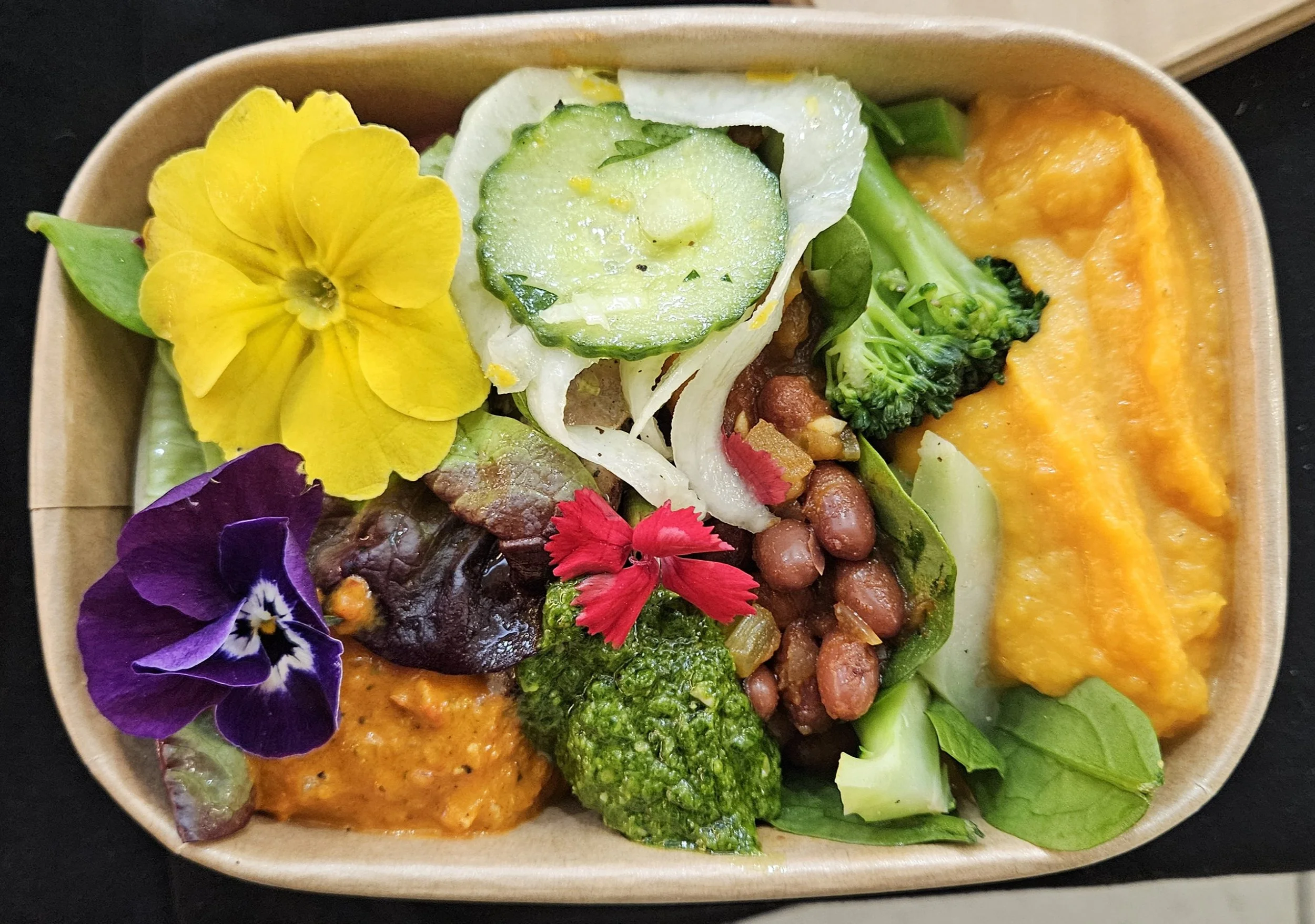 A bowl of assorted fresh vegetables and edible flowers, including cucumbers, broccoli, leafy greens, and purple and yellow flowers.