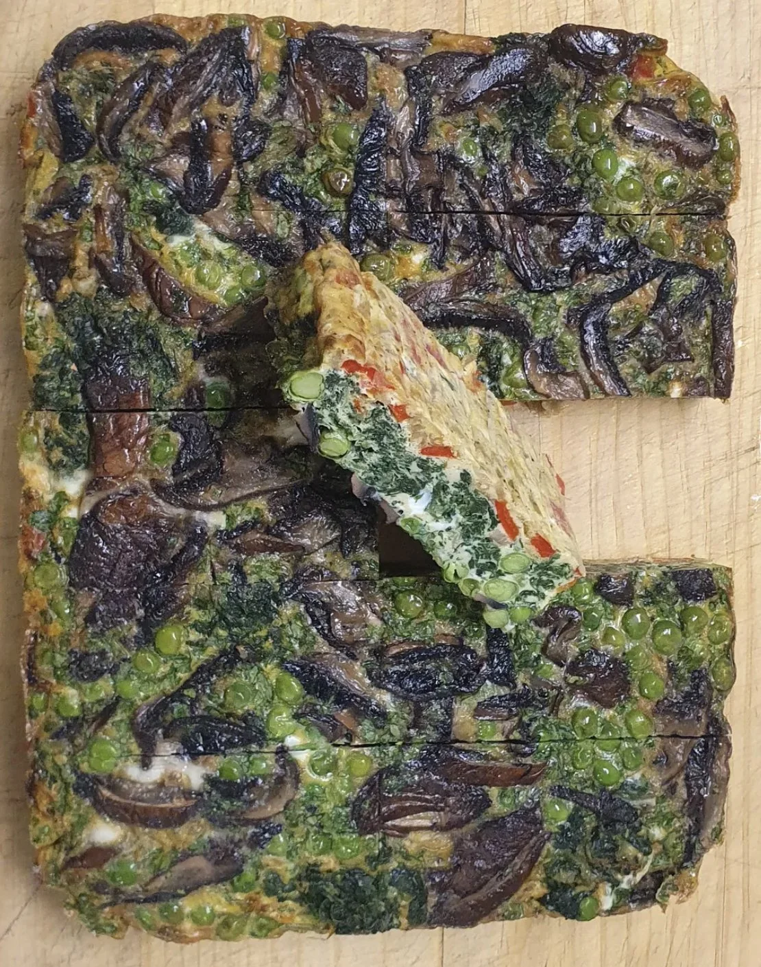 Vegetable frittata with mushrooms, green peas, and spinach, cut into squares and one piece slightly lifted, placed on a wooden surface.