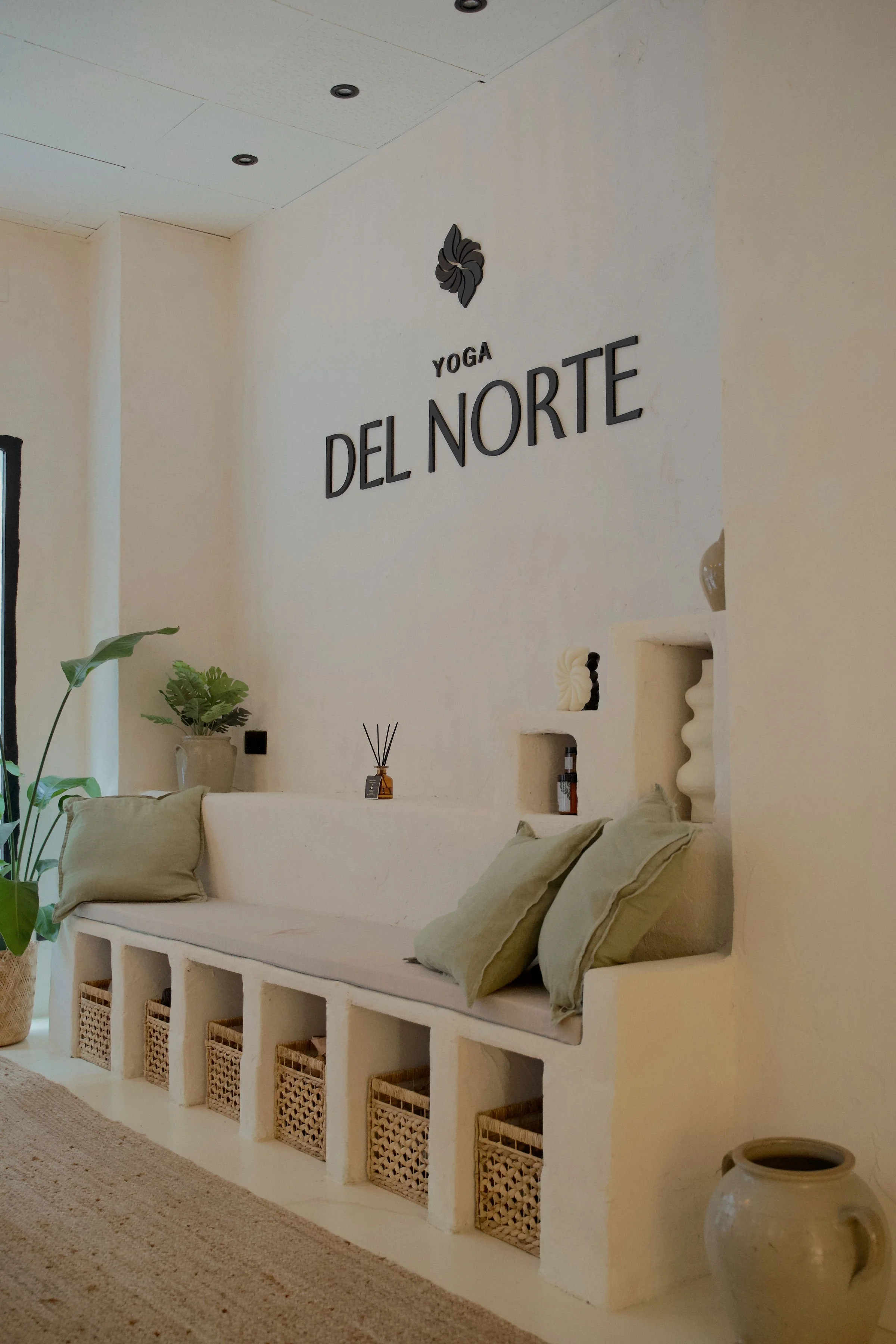 Interior de un espacio de yoga llamado 'Yoga del Norte' con un banco de concreto con cojines, plantas y decoraciones en tonos neutros.