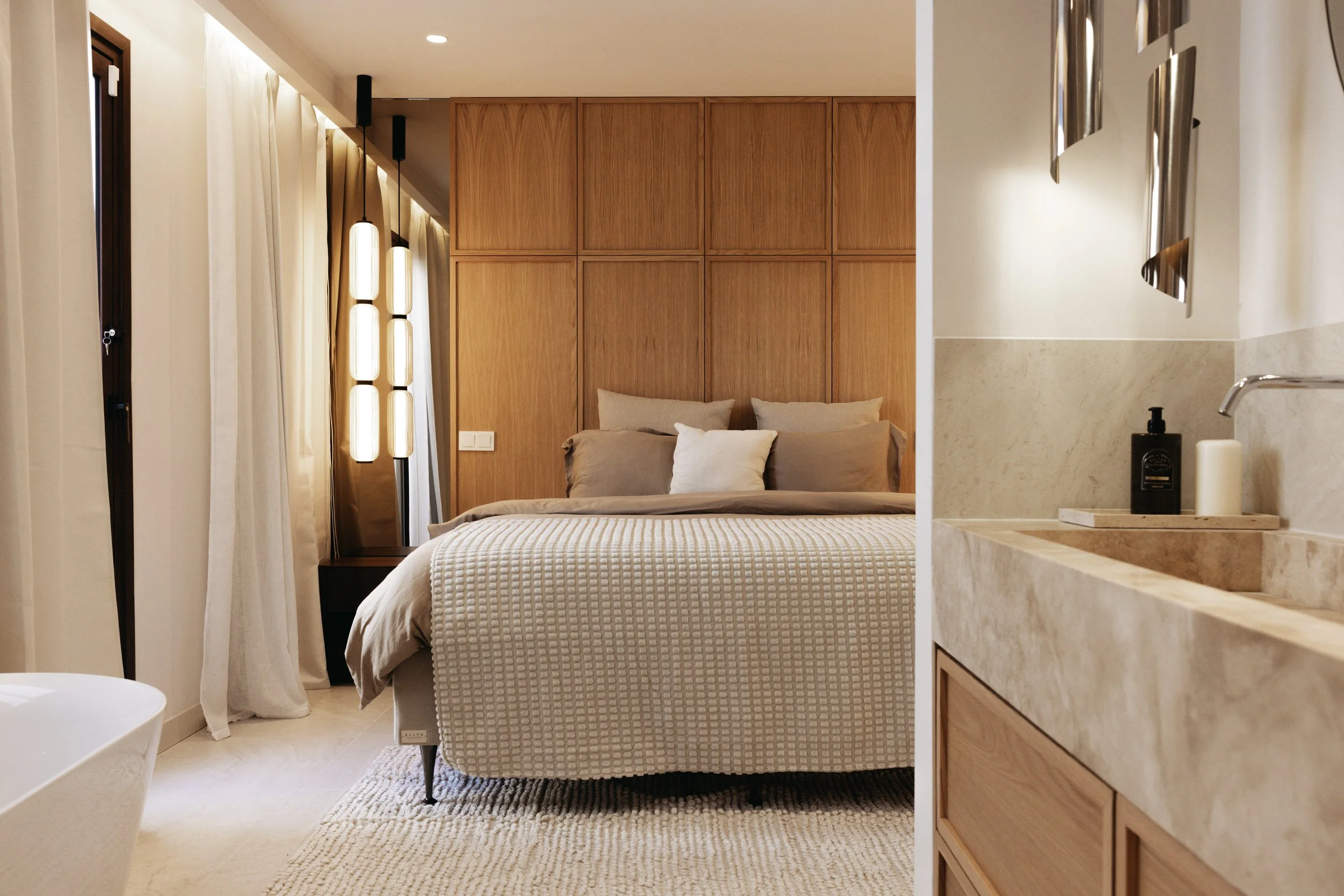 Sovrum med en bädd, träpanelväggar, utsikt mot en badrumssänkt med handtvätt och lysande lampor.