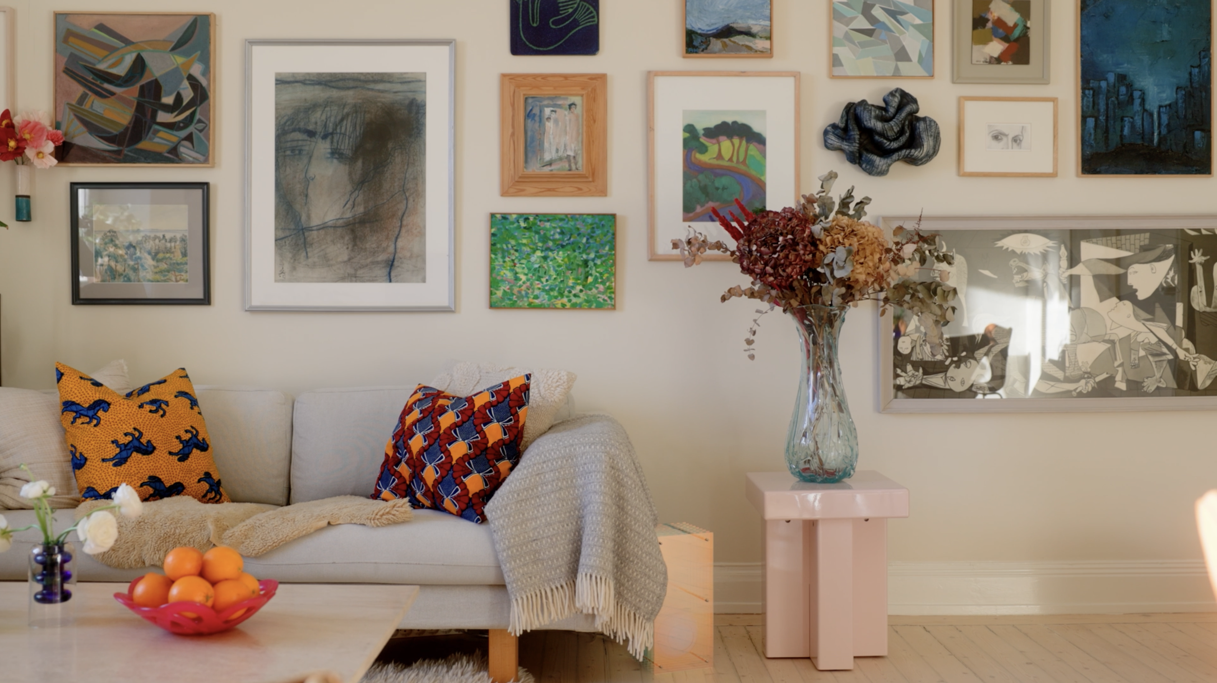 Vägg med flera konstverk, en soffa med kuddar, ett bord med en fruktskål och en stor vas med torkade blommor.