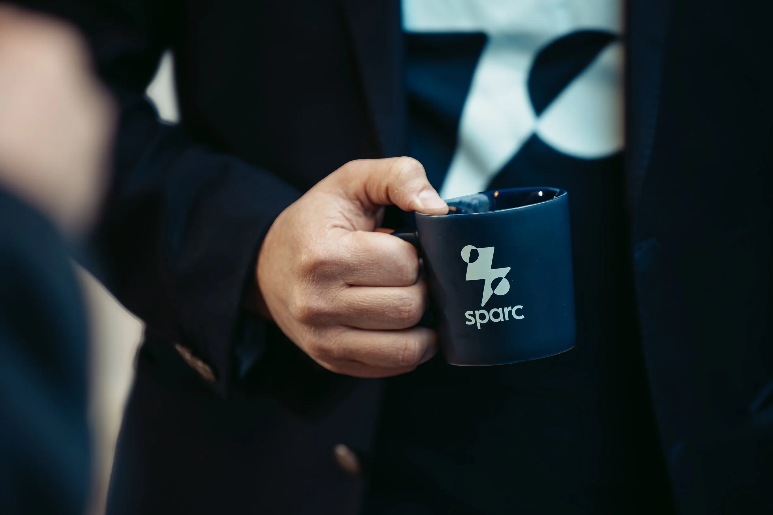 En person håller i en mörkblå kopp med logotypen 'sparc' på den, personens hand syns tydligt.