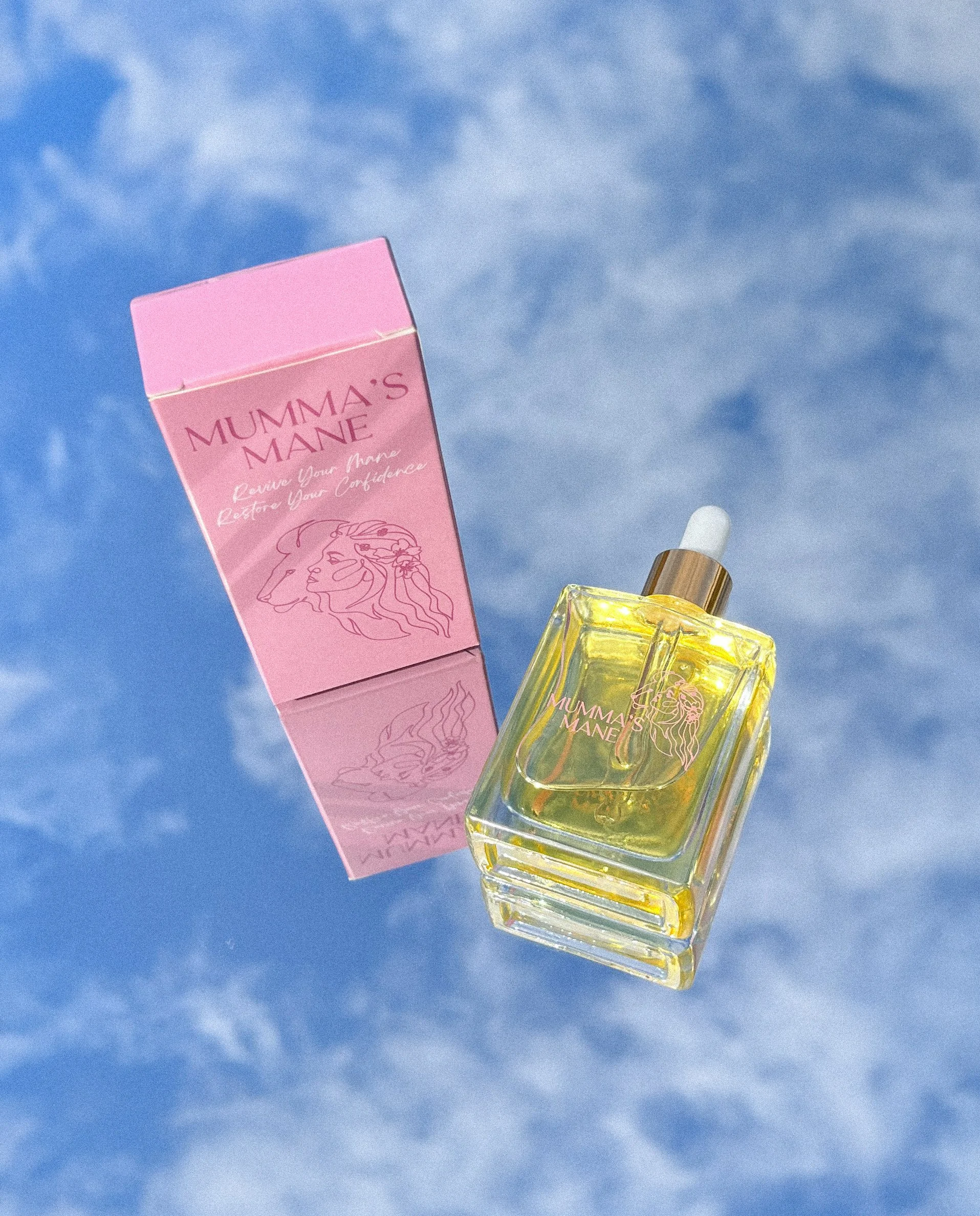 A bottle of perfume labeled 'Mumma's Mane' with a pink box beside it, set against a blue sky background.