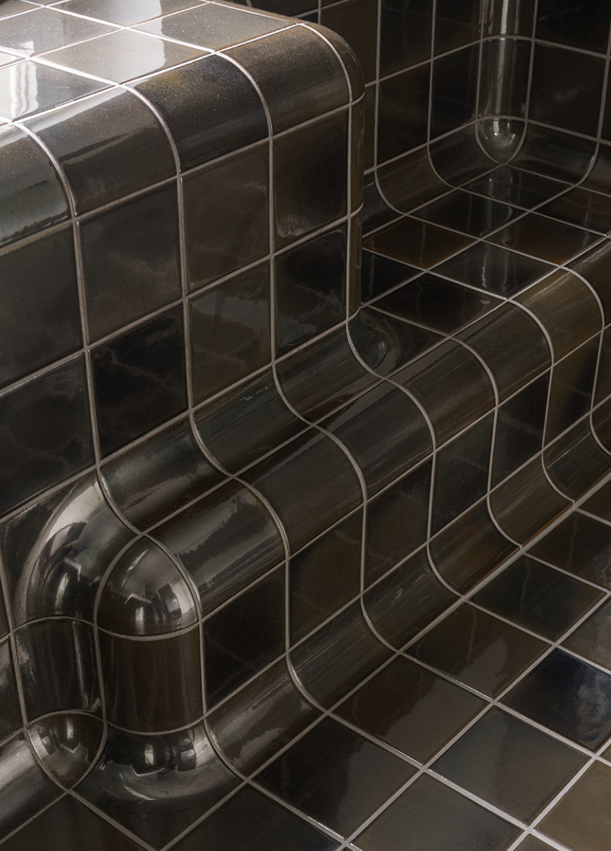 Glossy dark Japanese ceramic tiles with subtle tonal variation, wrapped over curved bench seating and rounded edges, showcasing smooth grout lines and sculptural detailing in a contemporary interior.