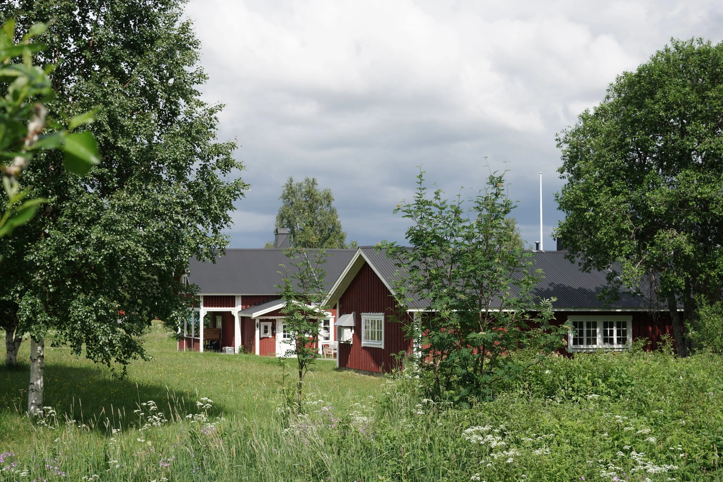 Ein traditionelles schwedisches rotes Holzhaus mit weißen Fenstern in einer grünen, ländlichen Umgebung unter einem bewölkten Himmel.