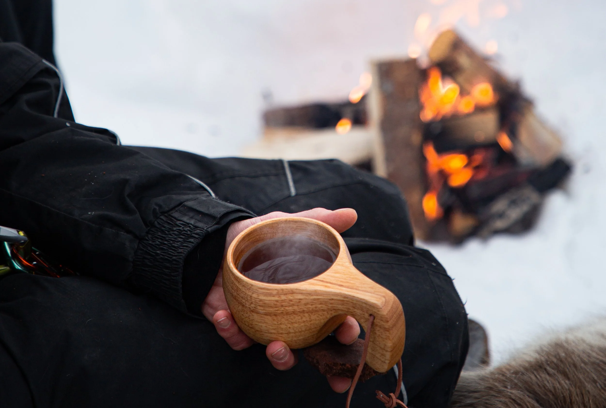 Eine Person hält eine Holzschale mit heißem Tee vor sich, im Hintergrund ein Feuer im Schnee.