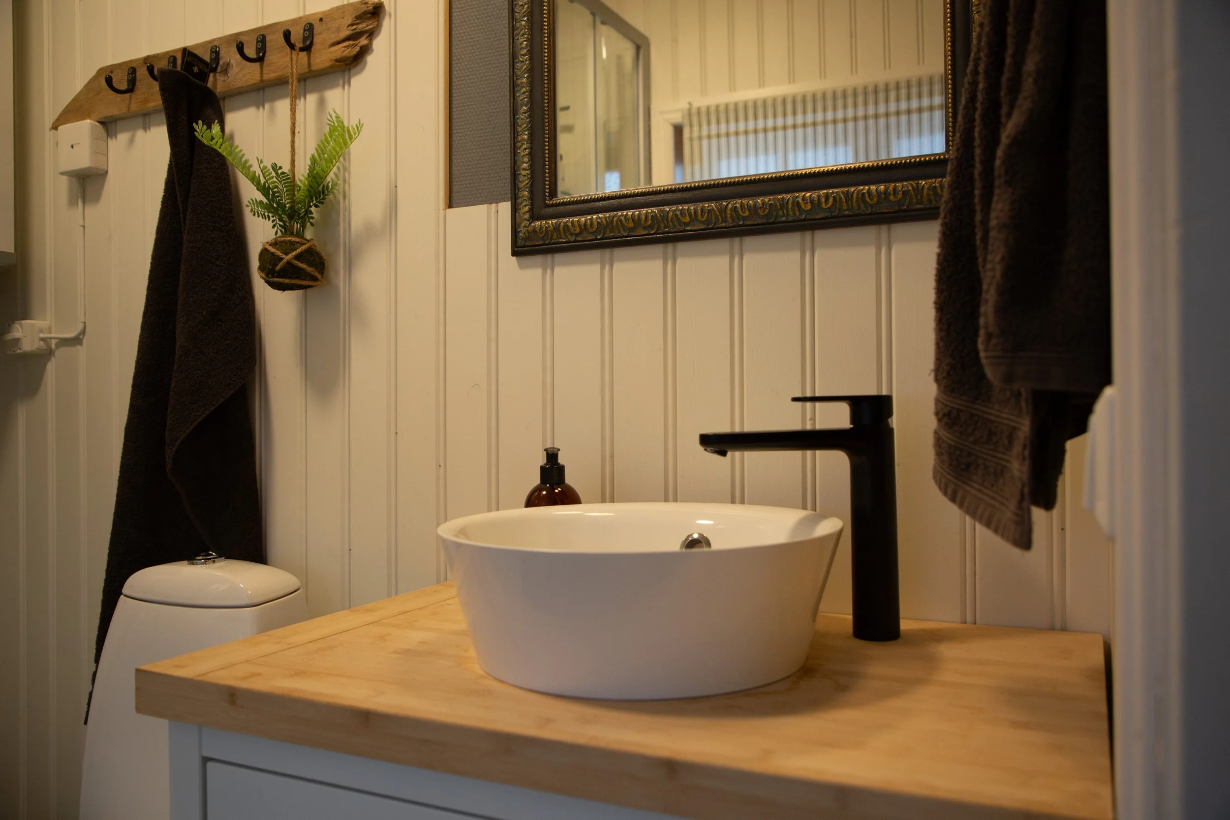 Badezimmer mit Vanity, schwarzer Wasserhahn, Spiegel, Handtücher und Pflanzen an der Wand.