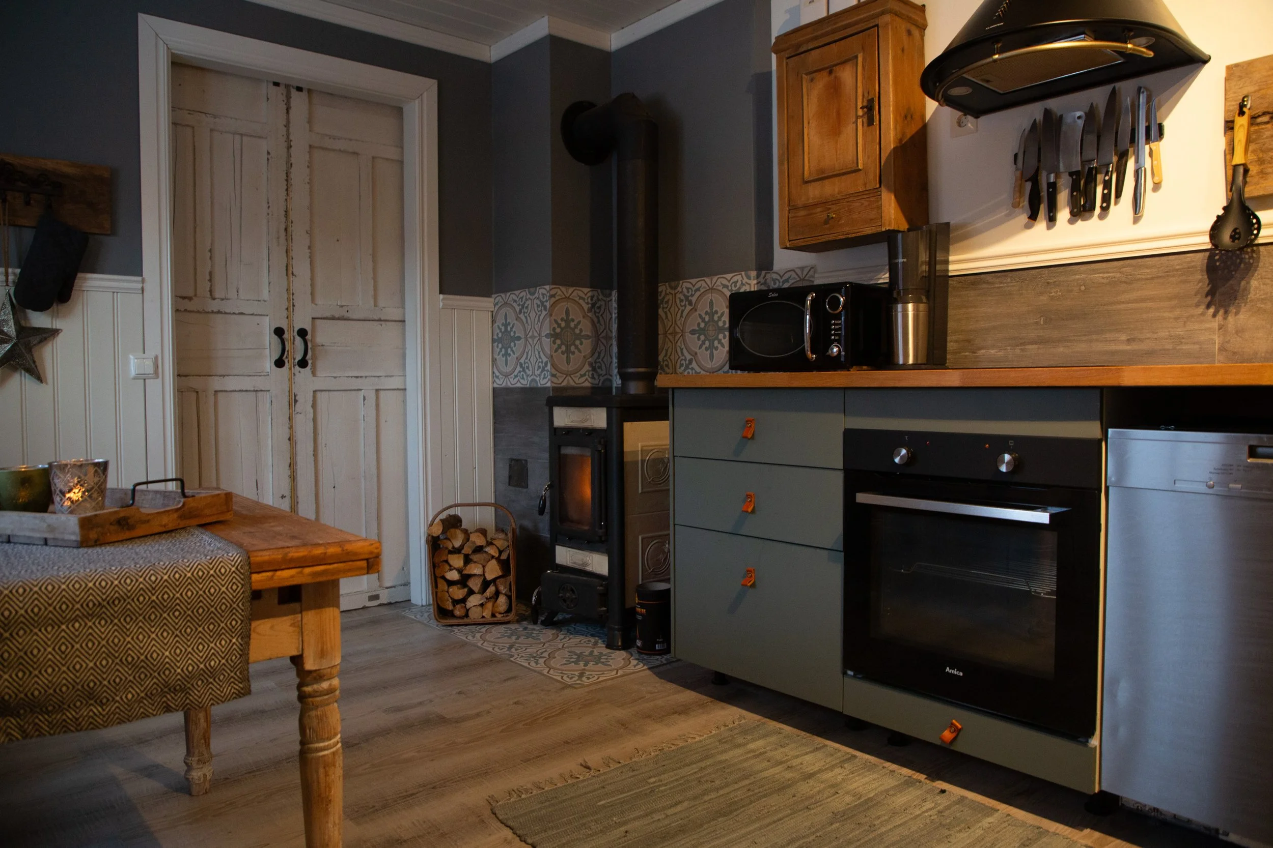 Küche mit Holzofen, Küchenkästen, Küchenutensilien und einem Tisch mit Tischdecke