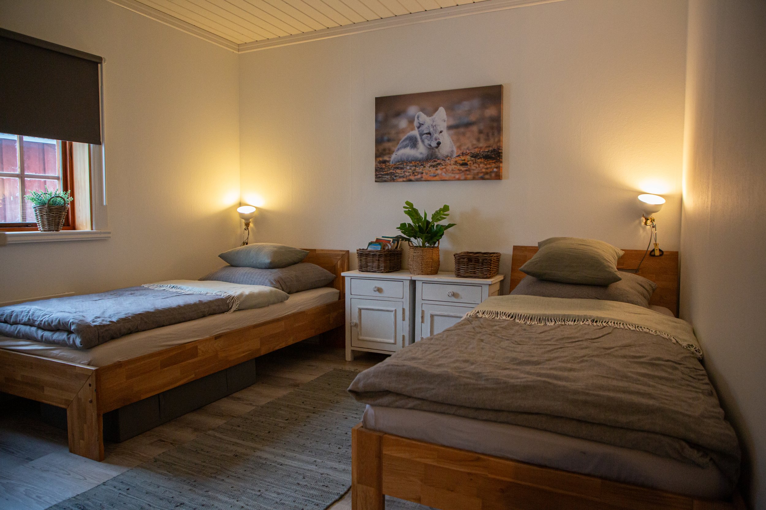 Gemütliches Schlafzimmer mit zwei Einzelbetten aus Holz, einer weißen Nachttisch mit Pflanzen und Korb, Wandbilder eines Wolfsjungen, seitliche Beleuchtung und einem Fenster mit Rollschatten.
