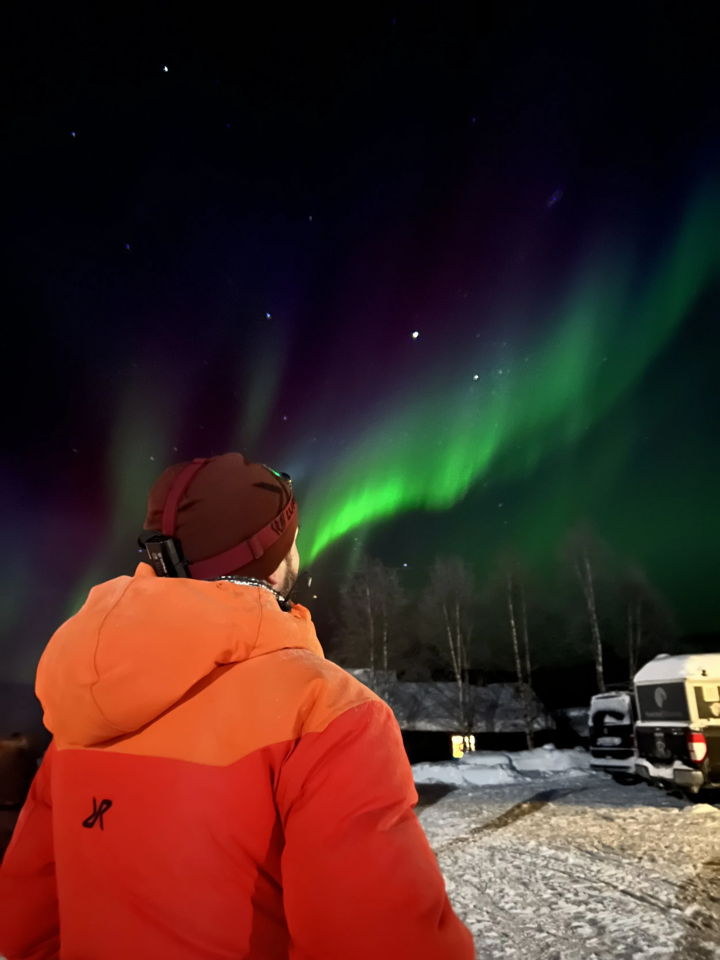 Ein Mann in orange Jacke und Mütze schaut in den Nachthimmel, wo grüne Nordlichter und Sterne sichtbar sind, in einer verschneiten Landschaf