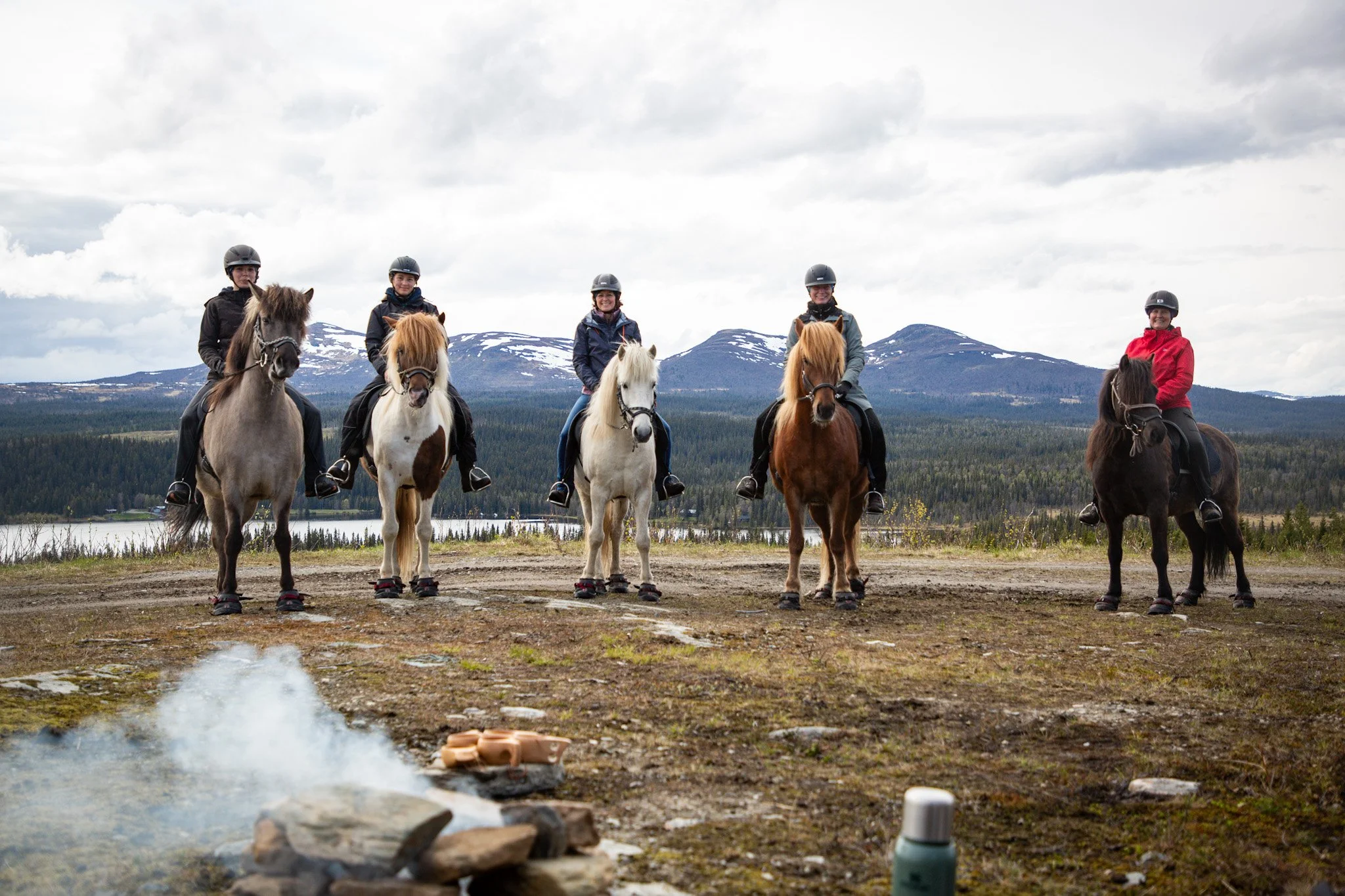Fünf Personen reiten auf Pferden in einer ländlichen Landschaft mit Bergen im Hintergrund.