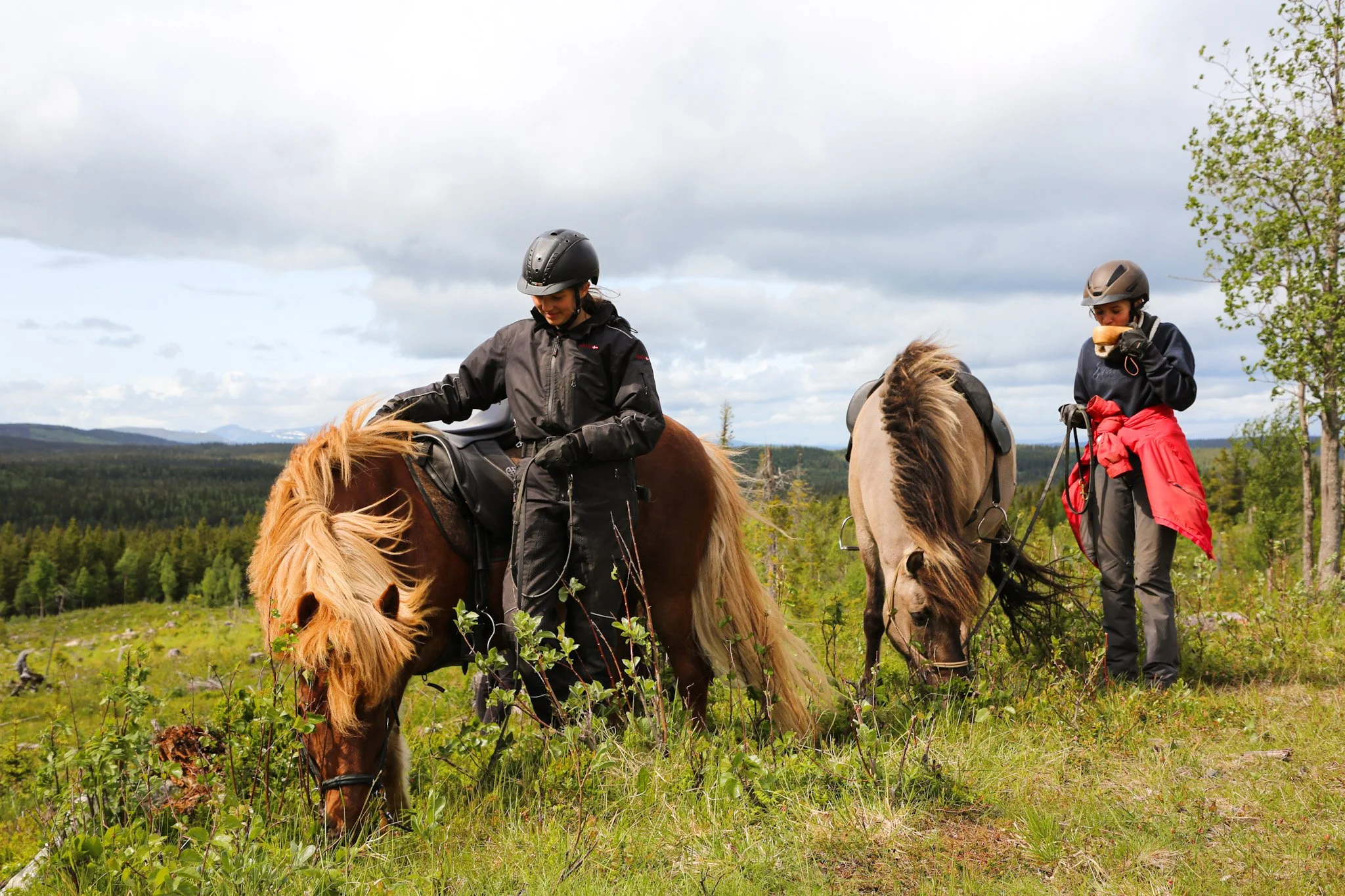 Zwei Frauen reiten auf Pferden im Grünen bei bewölktem Himmel, eine füttert die Pferde, die andere steht auf dem Boden.