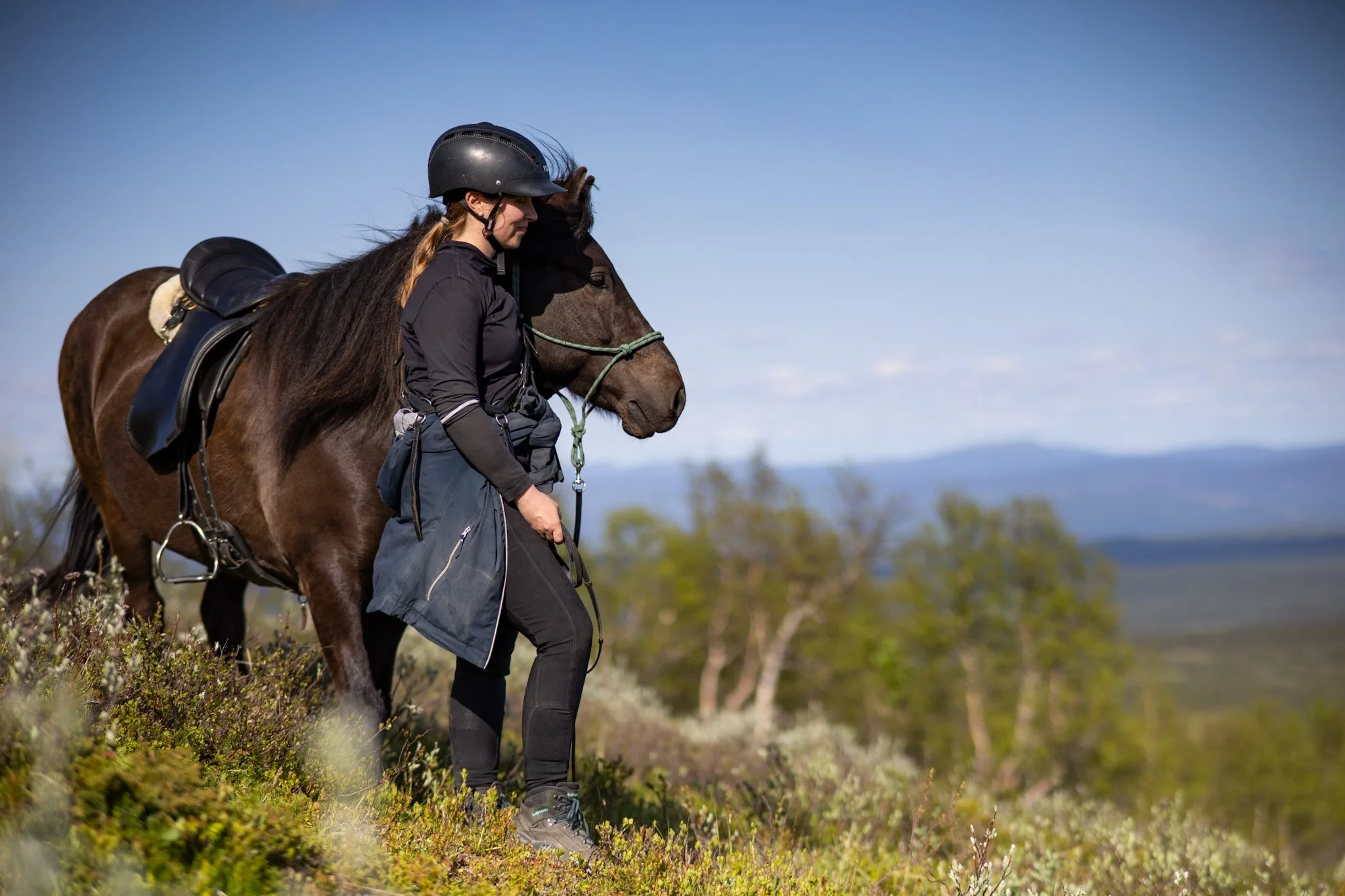 Eine Frau mit Reithelm und Reitbekleidung steht mit einem braunen Pferd in einer natürlichen Landschaft mit Bäumen und Bergen im Hintergrund.