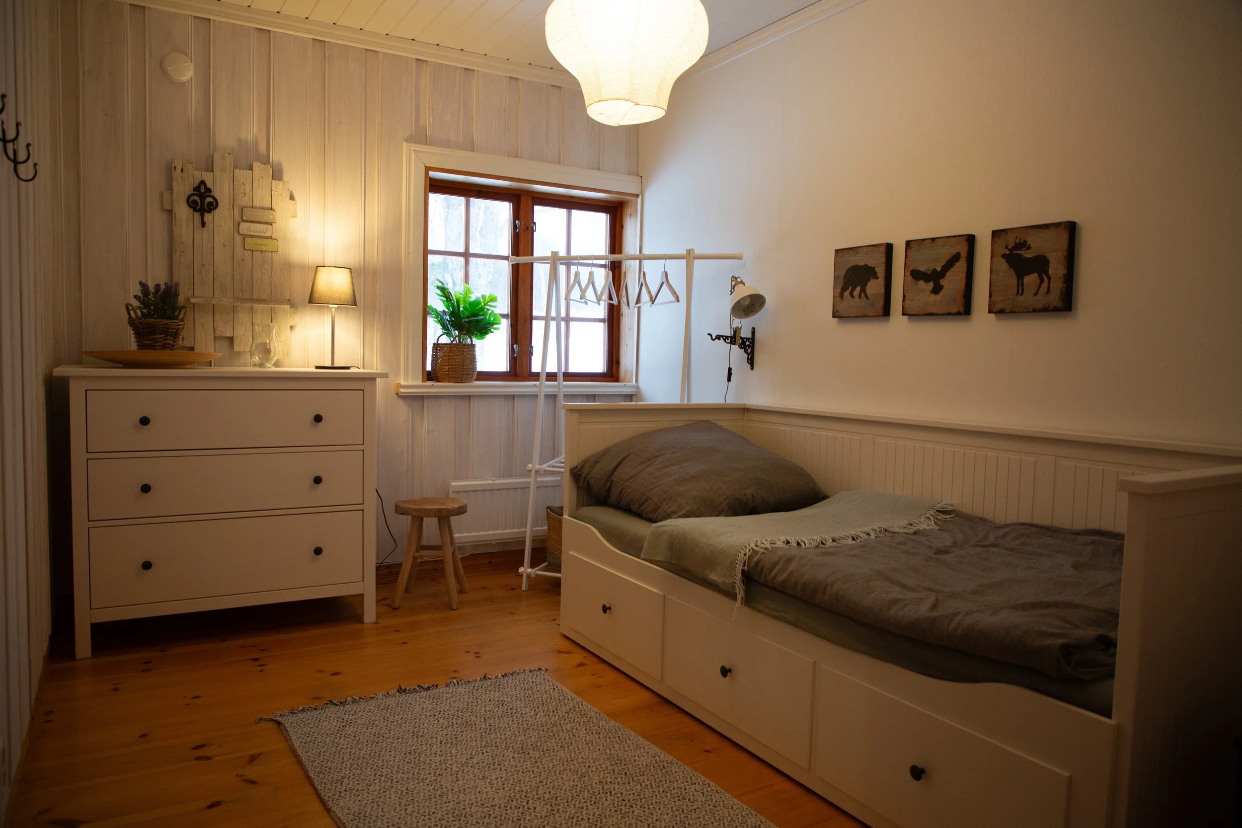 Gemütliches Schlafzimmer mit weißem Bett, Nachttisch, Holzlampe, Fenster mit Pflanzen, Wanddekoration und Holzfußboden.