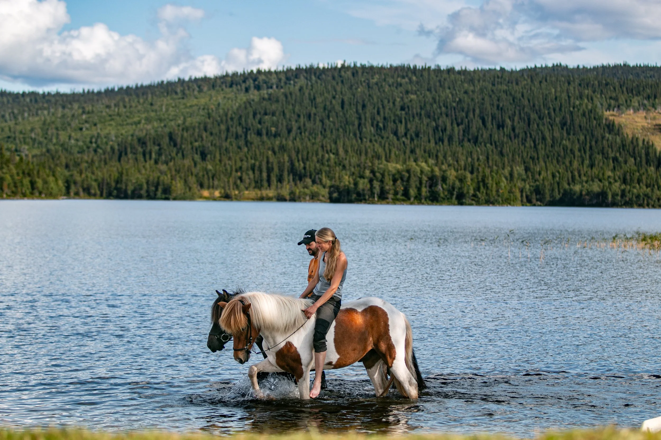 Zwei Menschen reiten barfuß auf einem Pony durch einen See vor einer bewaldeten Berglandschaft, bei sonnigem Himmel.