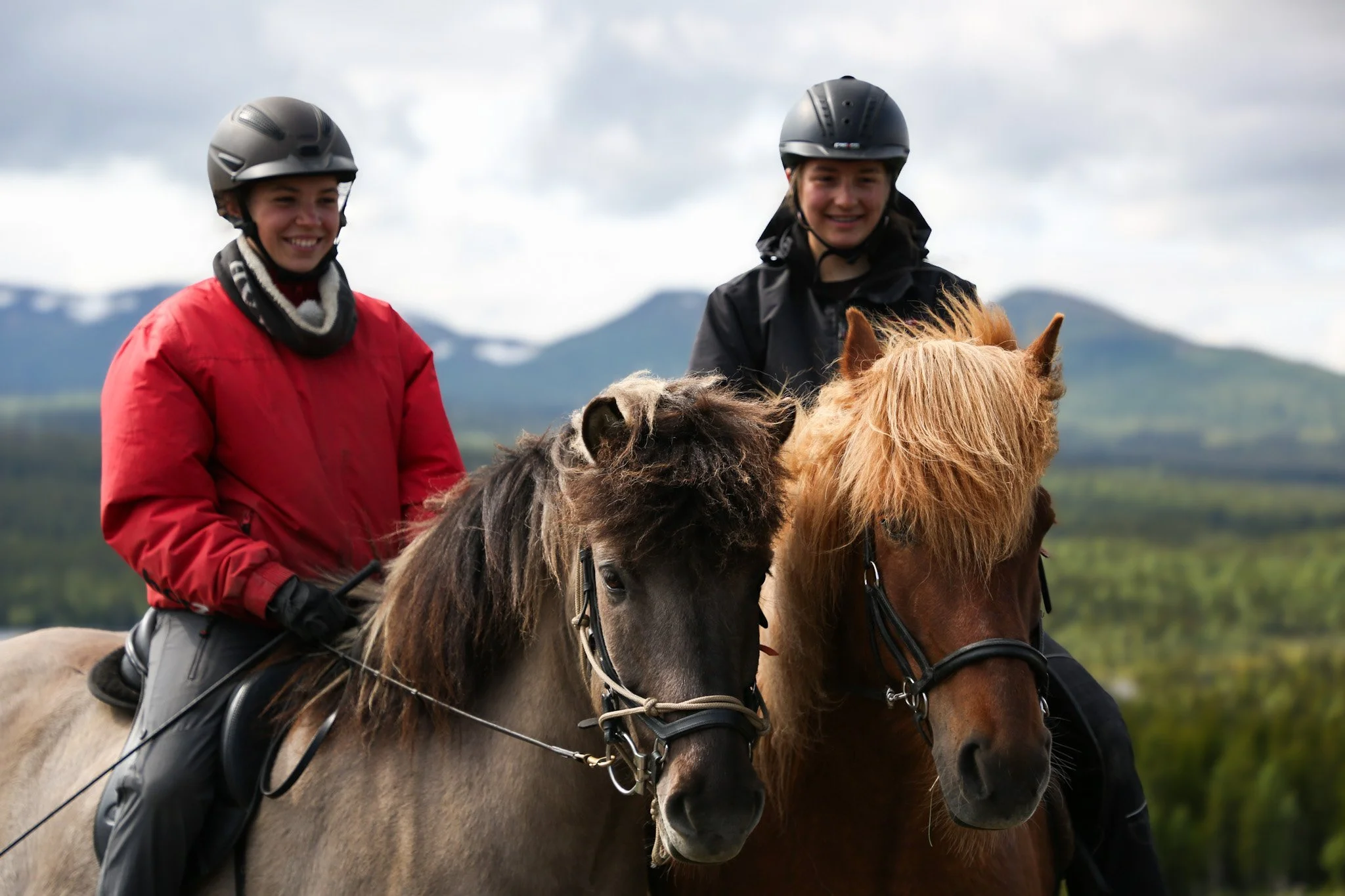 Zwei junge Frauen auf Pferden, beide tragen Helme, umgeben von einer Landschaft mit Bergen im Hintergrund.