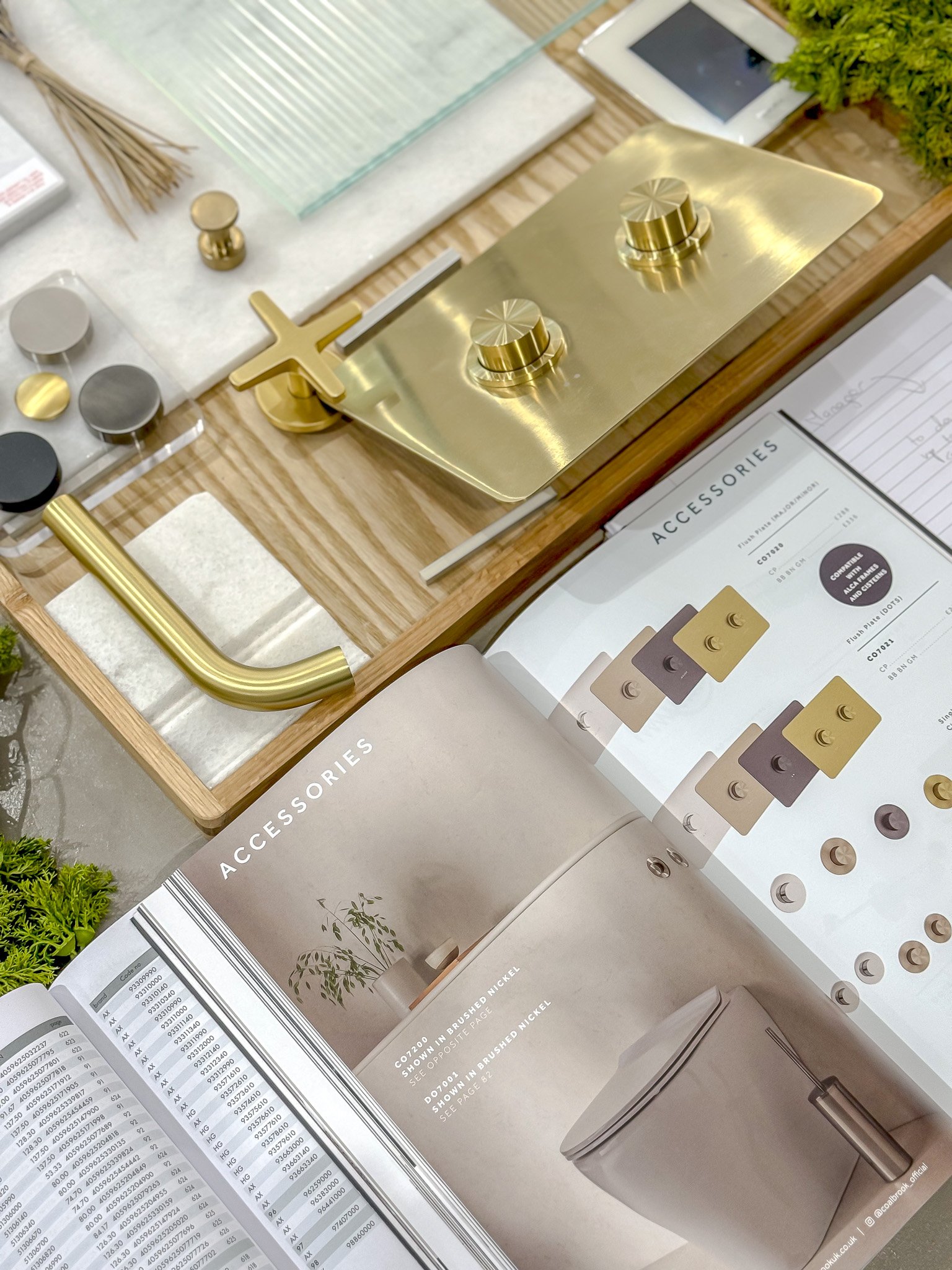Display table with various hardware and accessory samples, including a gold-colored kitchen faucet, a small metallic knob, and color swatches, along with printed catalogs and a small gray trash bin.