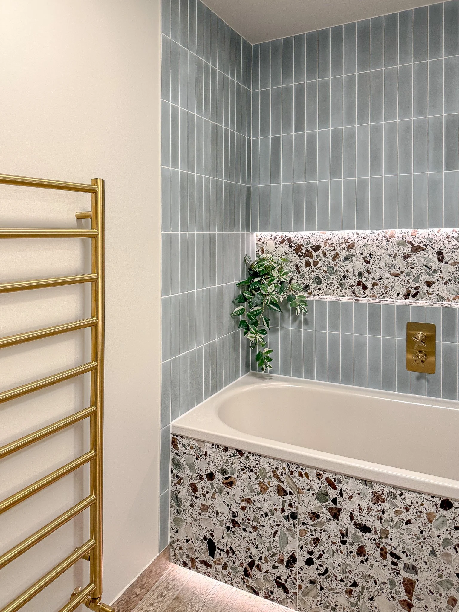 Contemporary bath with terrazzo tiled panel, soft grey vertical wall tiles, brushed brass bath filler and matching heated towel rail in a bespoke luxury bathroom